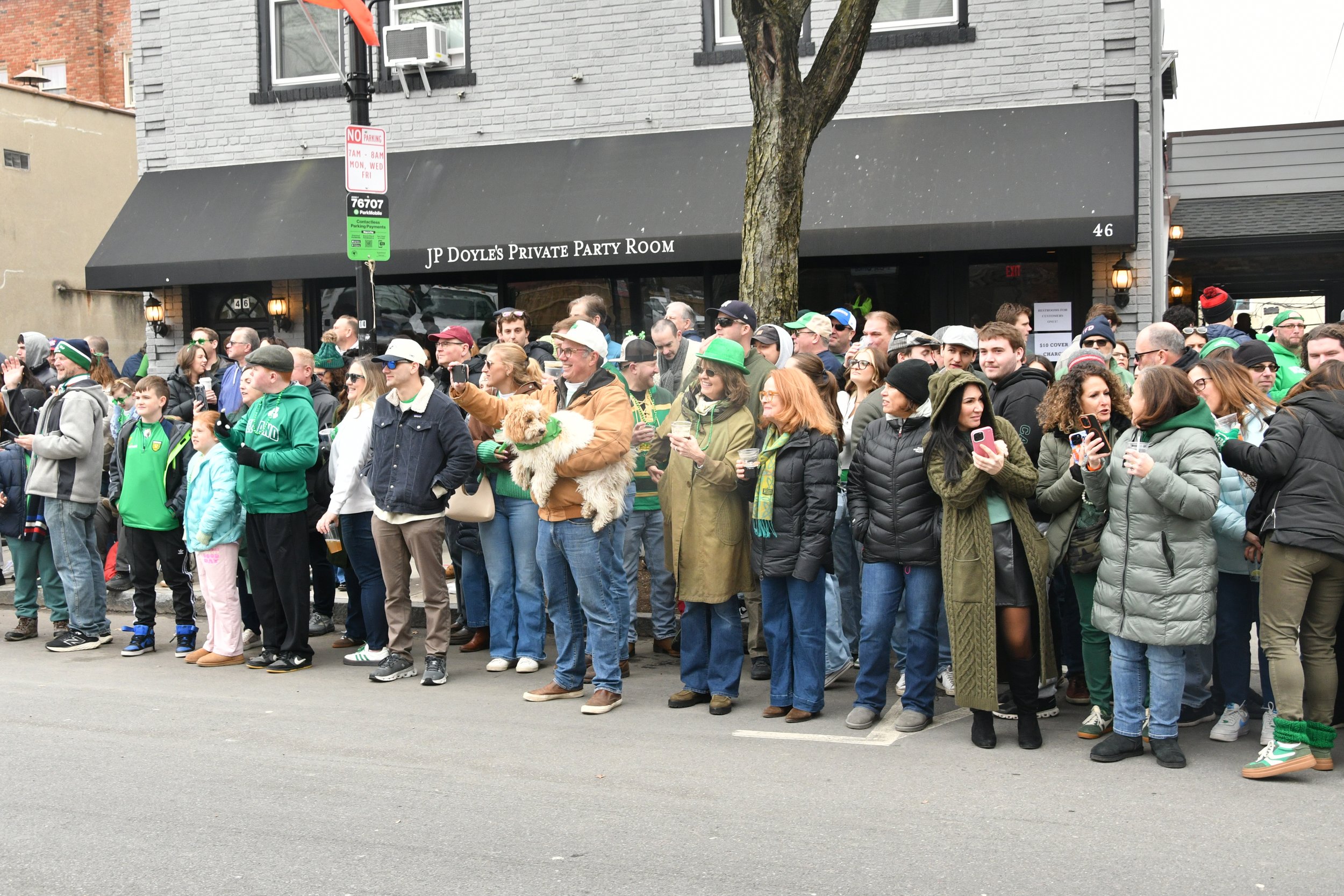 A large group of people standing on the sidewalk in front of a grey brick building with a sign reading "JP Doyle's Private Party Room." The crowd is festive, with many wearing green hats, scarves, jackets, and apparel, and some holding drinks. The we