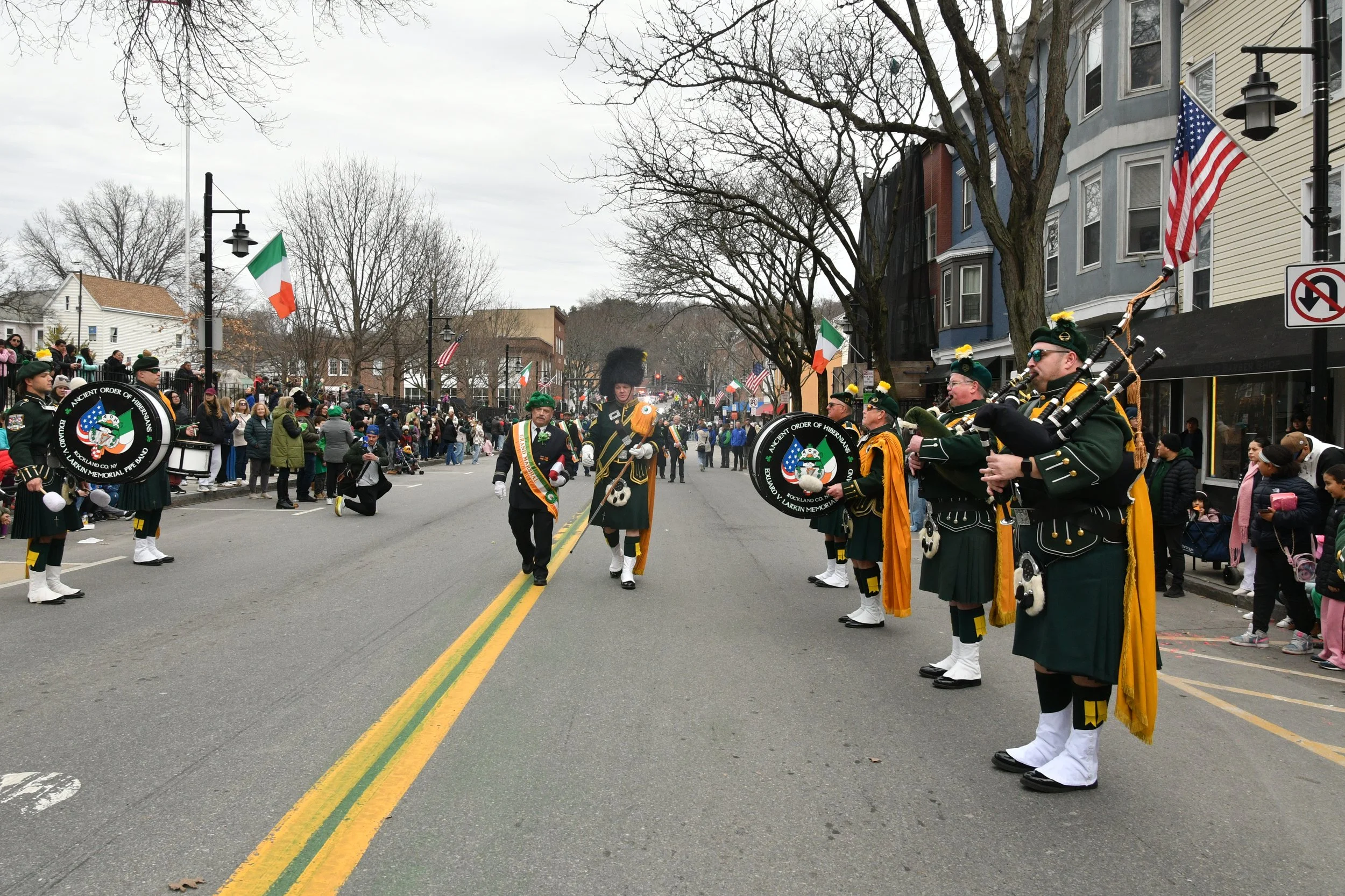 A parade with bagpipers in green uniforms and hats marching down a city street, lined with spectators and decorated with flags, including Irish flags and American flags, on a cloudy day.
