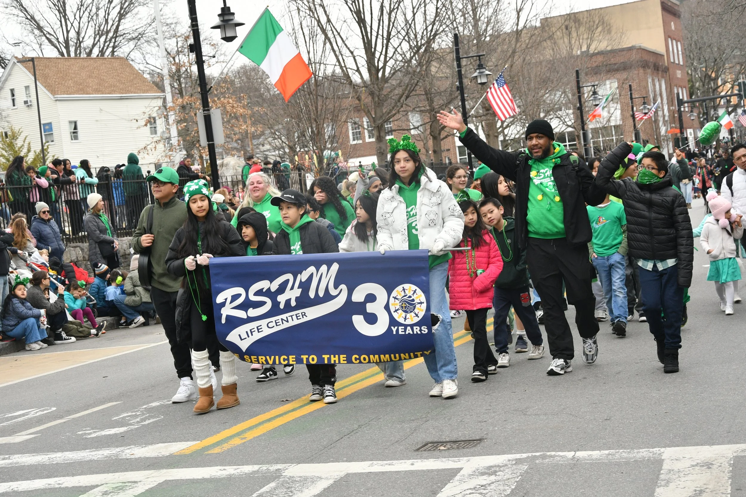 A parade with a group of people walking down the street, holding a blue banner that reads 'RSYM 30 Years Service to the Community.' Participants and onlookers are dressed warmly, many wearing green clothing and St. Patrick's Day accessories. Flags, i