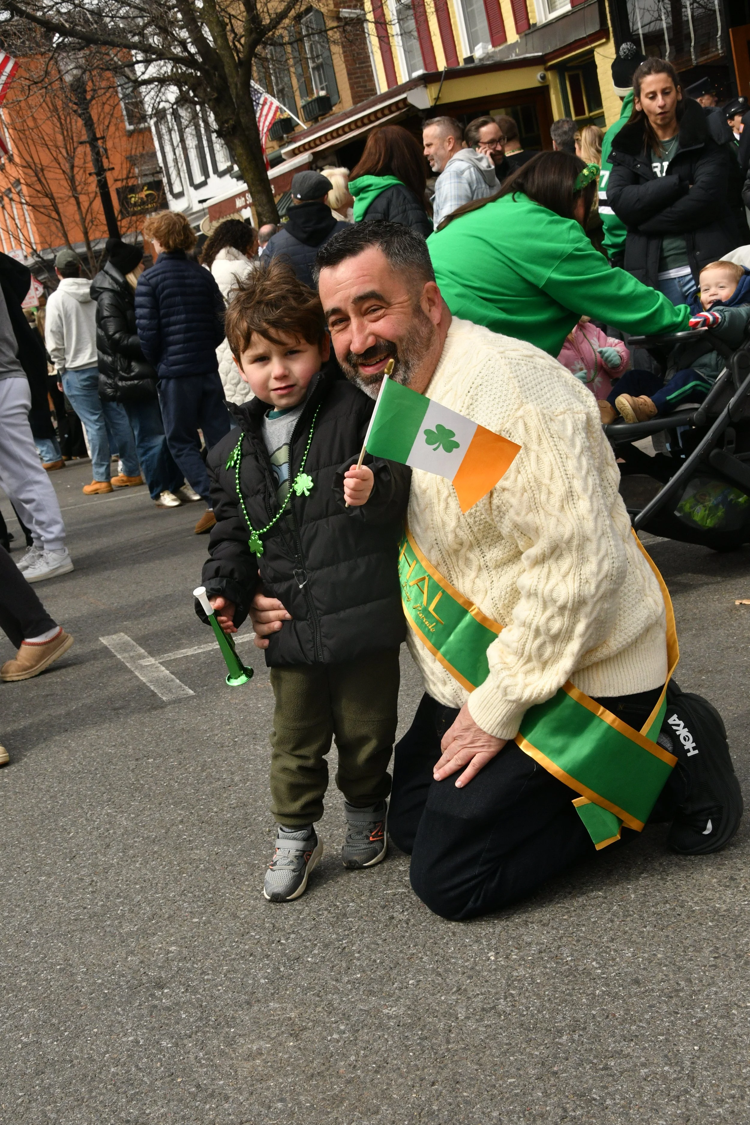 A man and a boy celebrating St. Patrick's Day outdoors. The man is kneeling on one knee, wearing a green sash and sweater, while the boy is holding a small Irish flag and wearing a black jacket with green bead necklaces. The background shows a crowd 