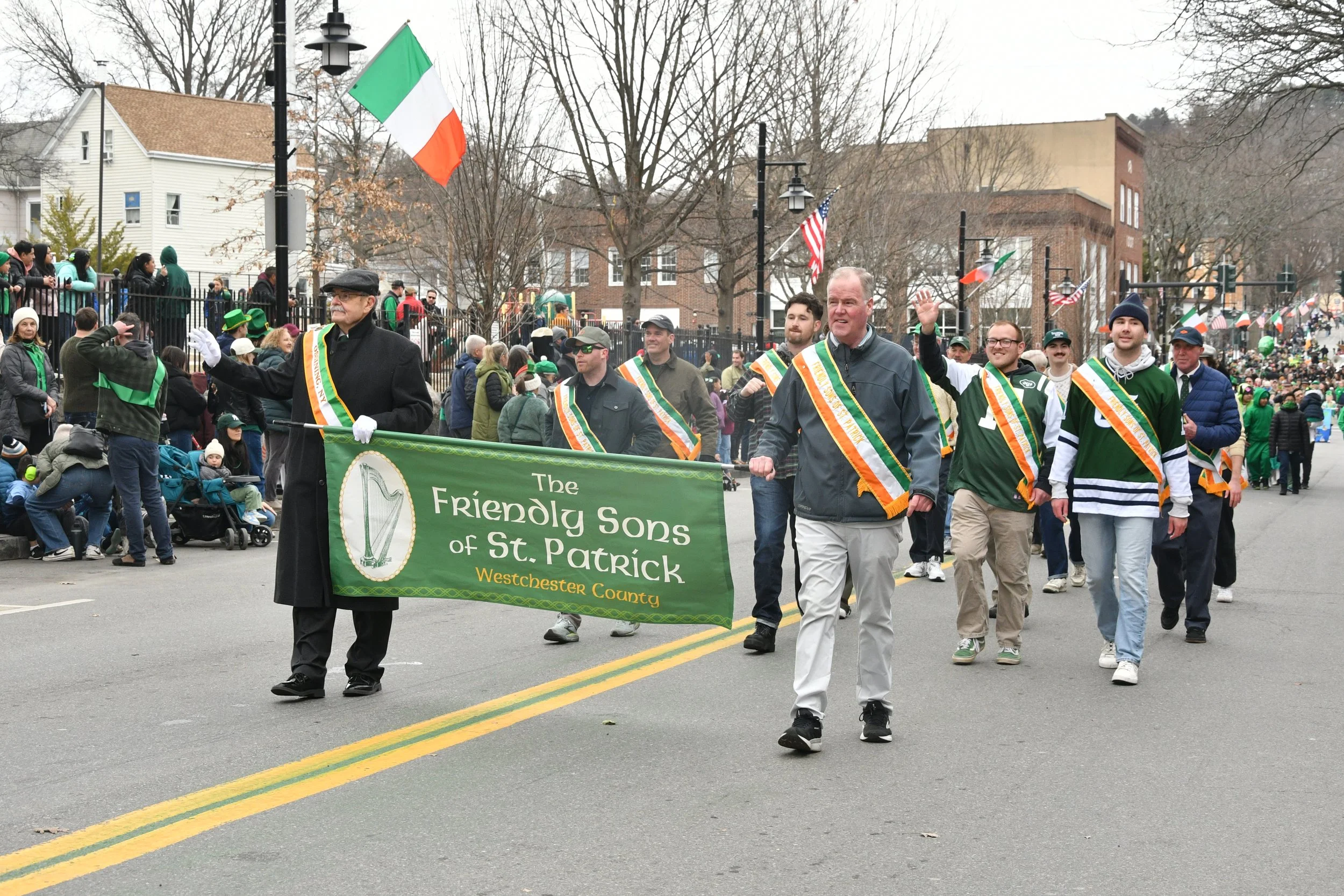 A parade with people marching down a street, holding a green banner that reads 'The Friendly Sons of St. Patrick, Westchester County.' Participants are dressed in green and other casual clothing, some wearing sashes and hats, with American and Irish 
