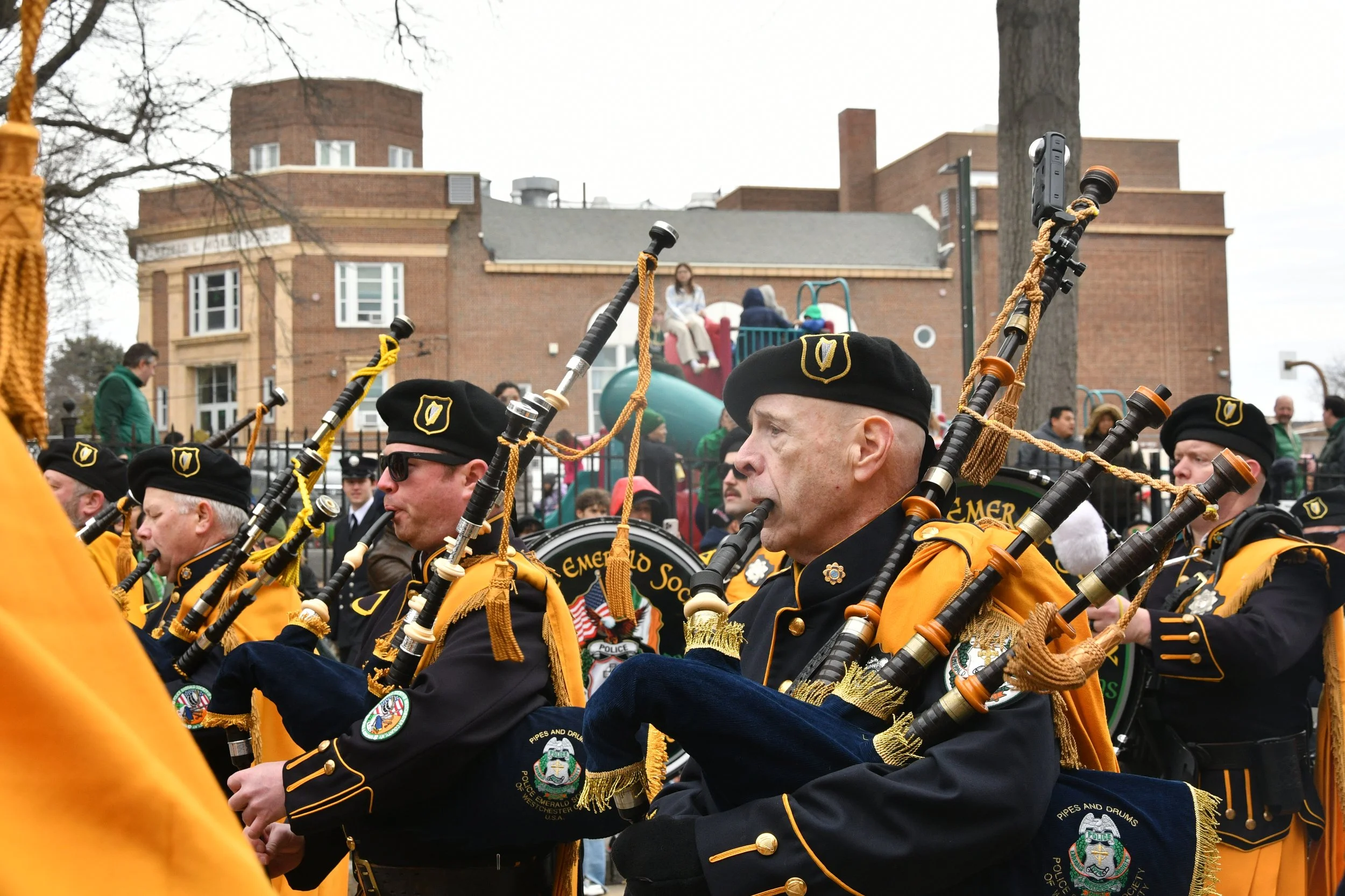 A parade featuring members of a police pipe band playing bagpipes. The band members are dressed in black uniforms with yellow accents, and some are wearing black berets with a shield patch. They are marching down a street with a brick building and a 