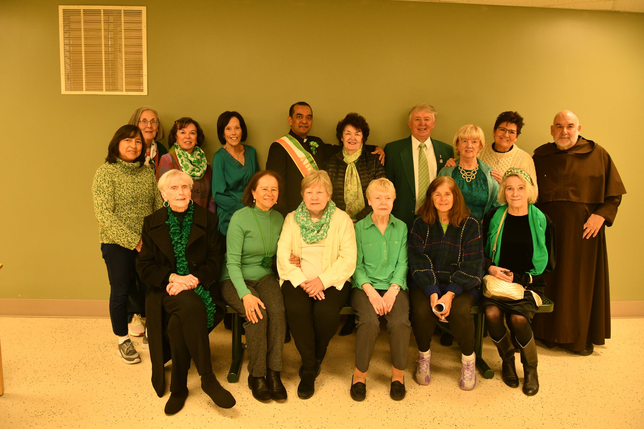 Group of 16 people, mostly women, gathered together indoors against a green wall, some sitting and some standing, wearing green clothing and accessories, celebrating St. Patrick's Day. One man wears a presidents sash, and another a brown religious ha