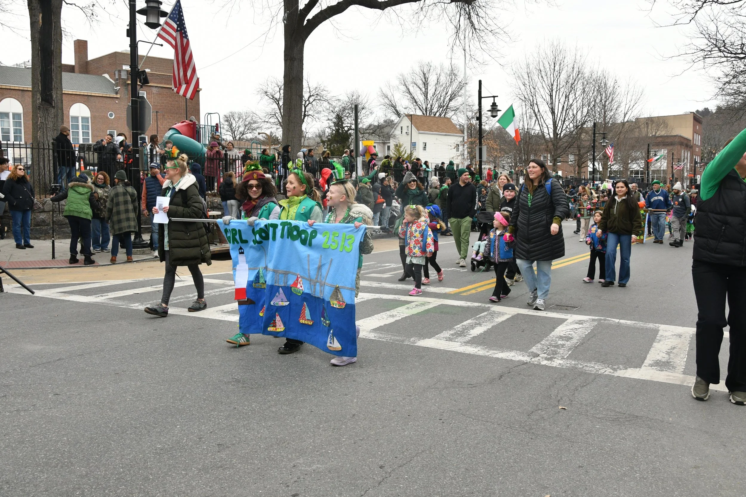 People participating in a parade on a city street, including children holding a colorful banner and various onlookers, with a playground and trees in the background.