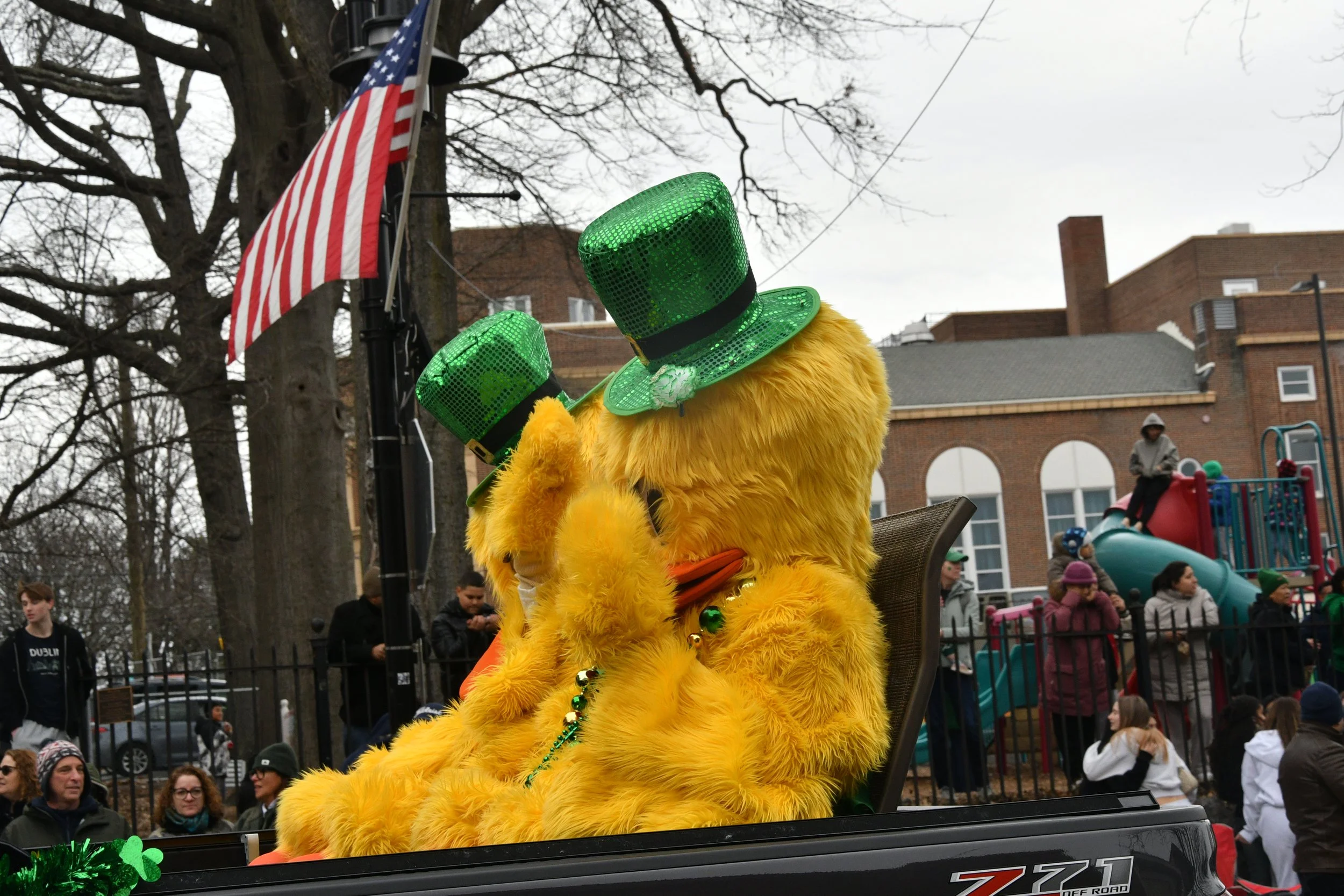 A person dressed as a yellow bird with large green glittery hats during a parade, with onlookers and a playground in the background.