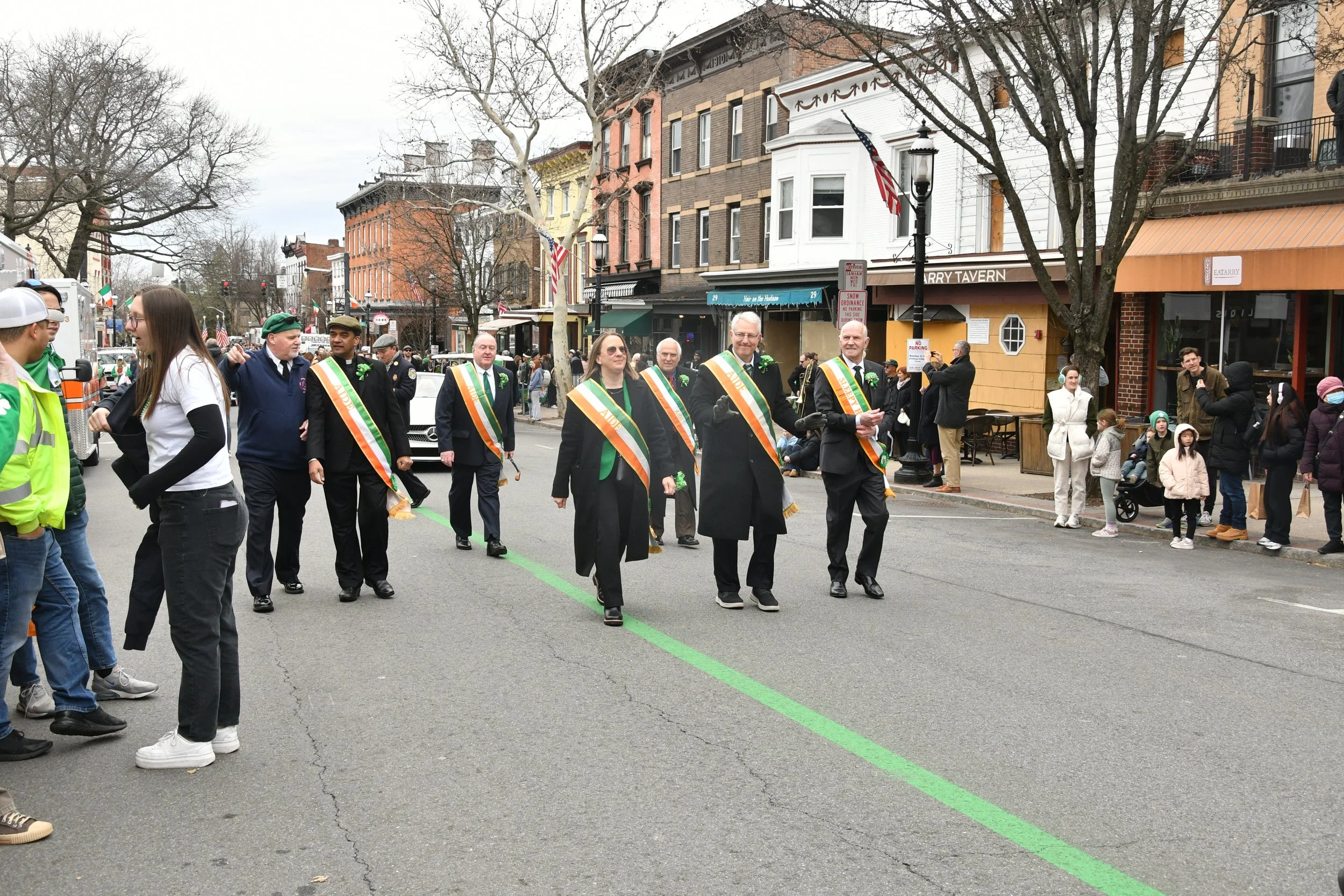 People participating in a parade on a city street, wearing sashes and coats, with some waving and others talking. Spectators are on the sidewalk watching the event.