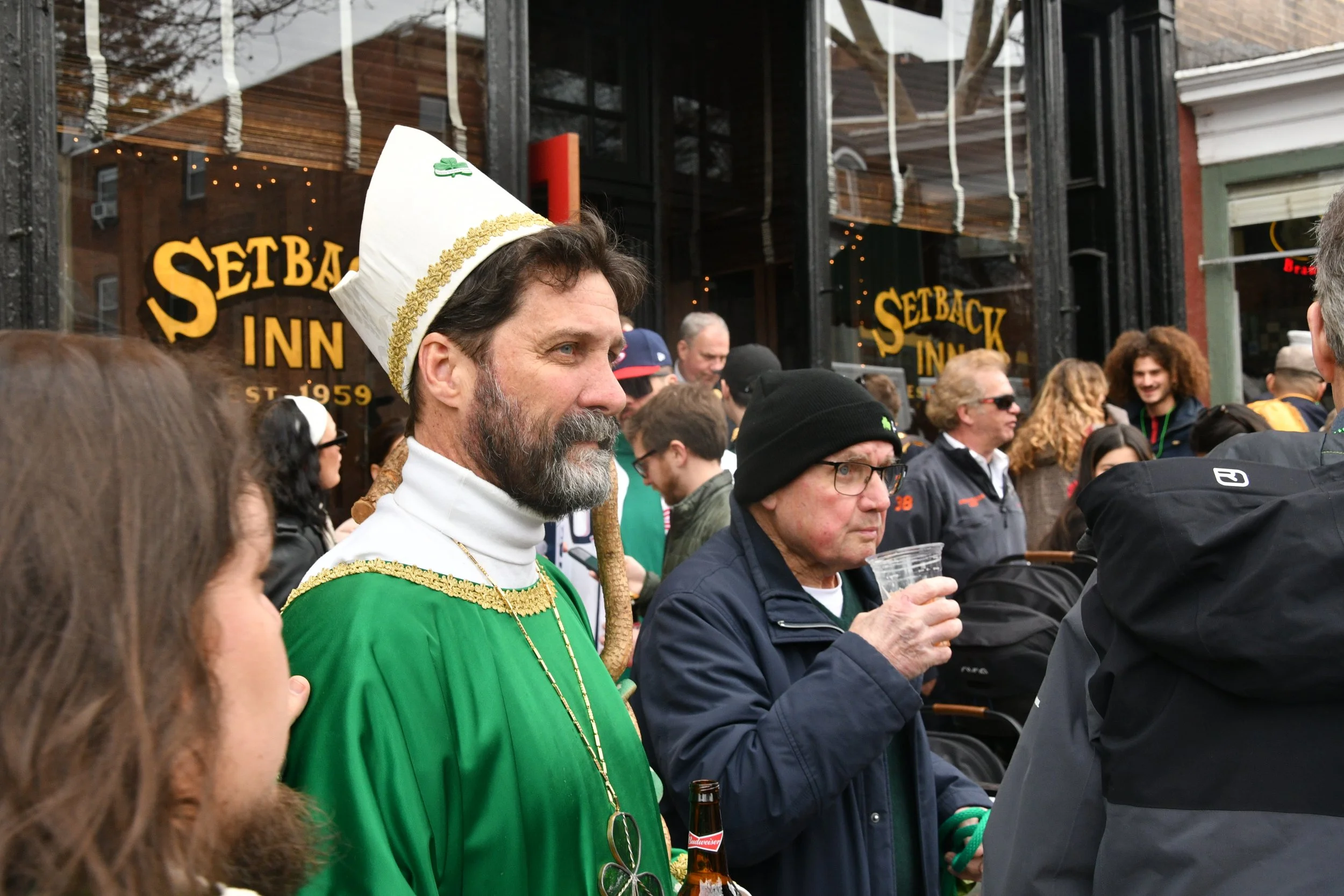 A crowd of people gathered outside a restaurant called Setback Inn, with one man in costume wearing a white hat with a clover and gold trim, dressed in green, holding a beer bottle.