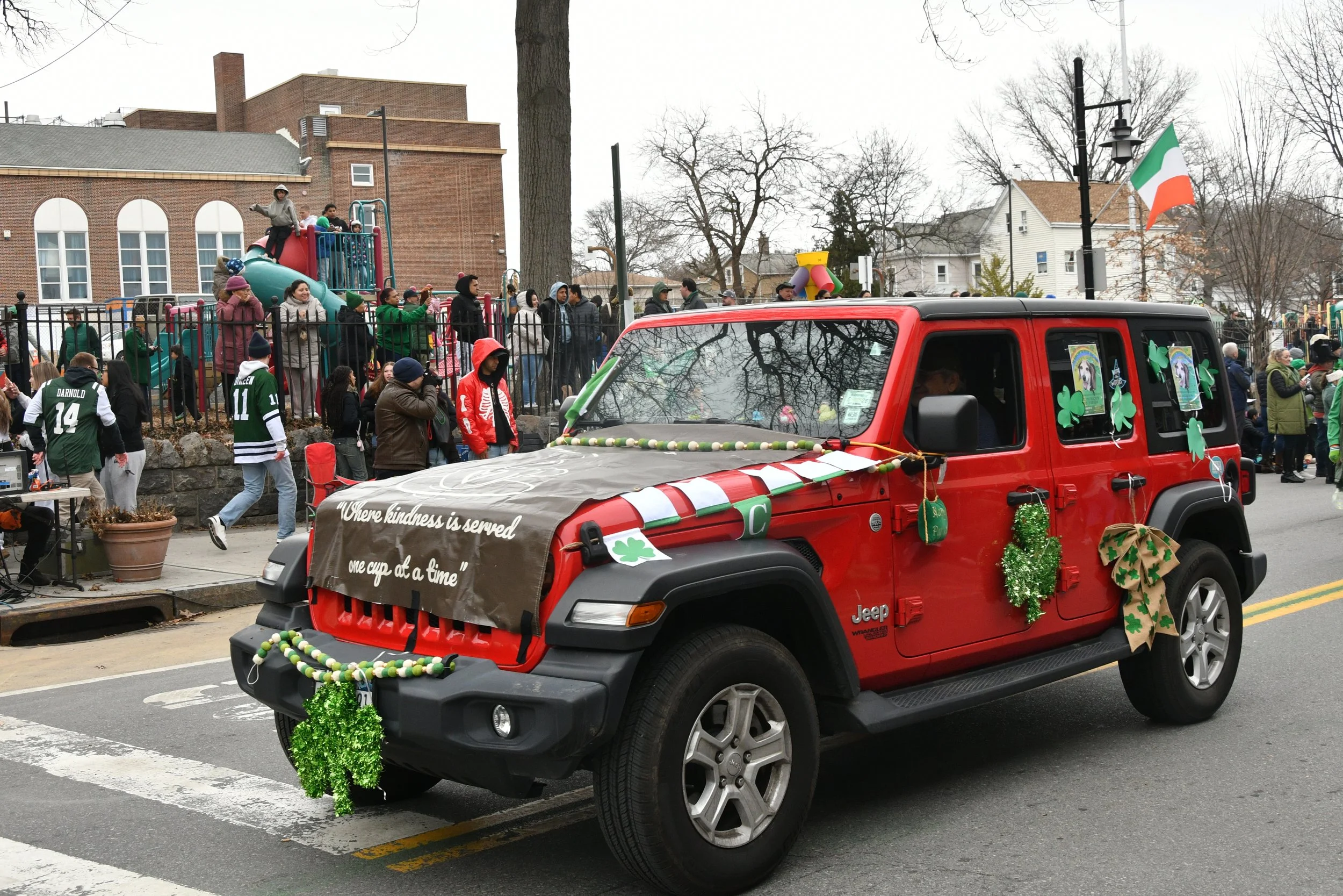 A parade float decorated with shamrocks and green and gold accents for St. Patrick's Day on a city street. People are gathered around, some dressed in green, and some watching from behind a fence. A red Jeep with St. Patrick's Day decorations is in t