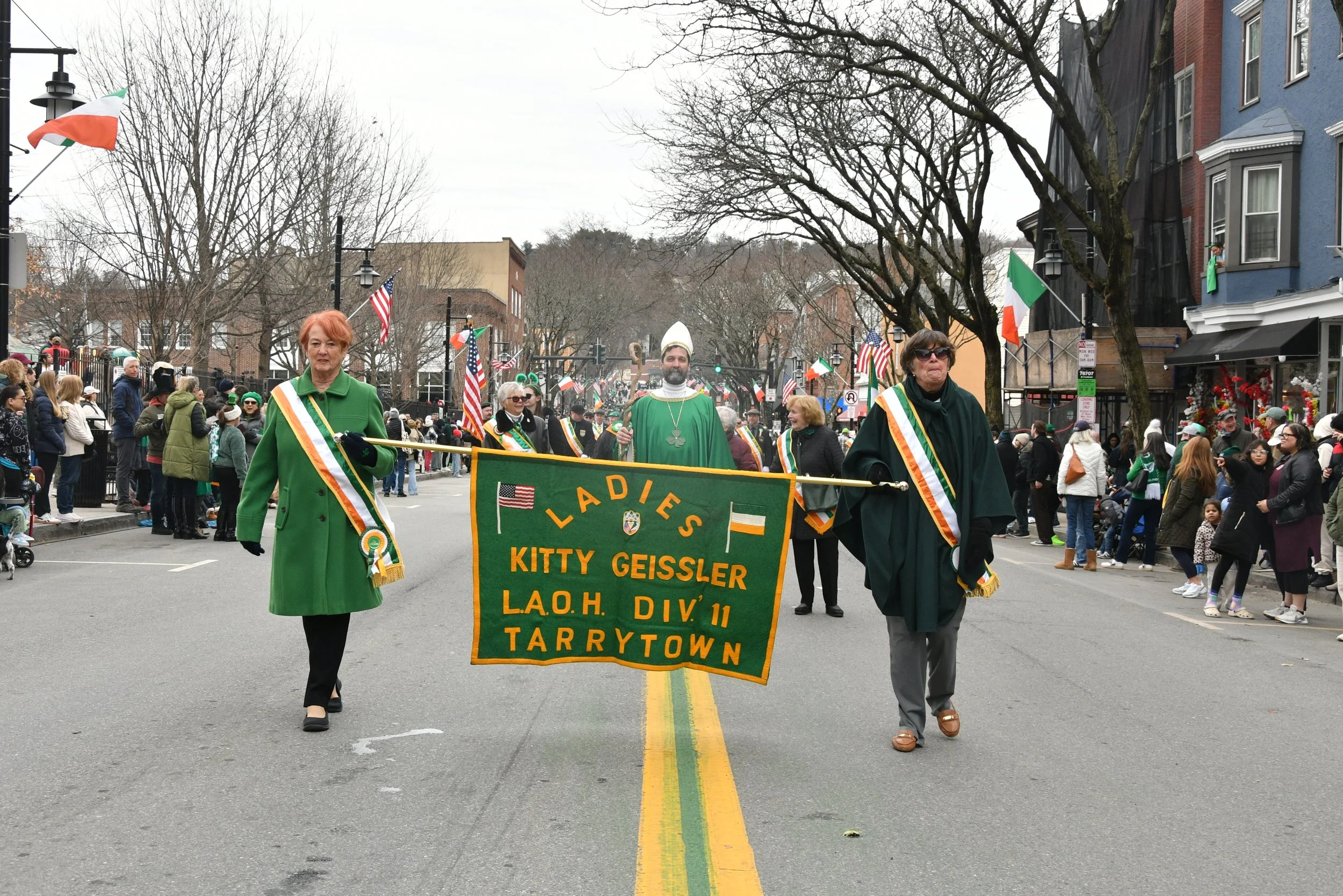 A parade with people marching down a street decorated with Irish flags. Front row includes women dress in green with sashes, holding a banner for Tarrytown, with spectators on the sides of the street.