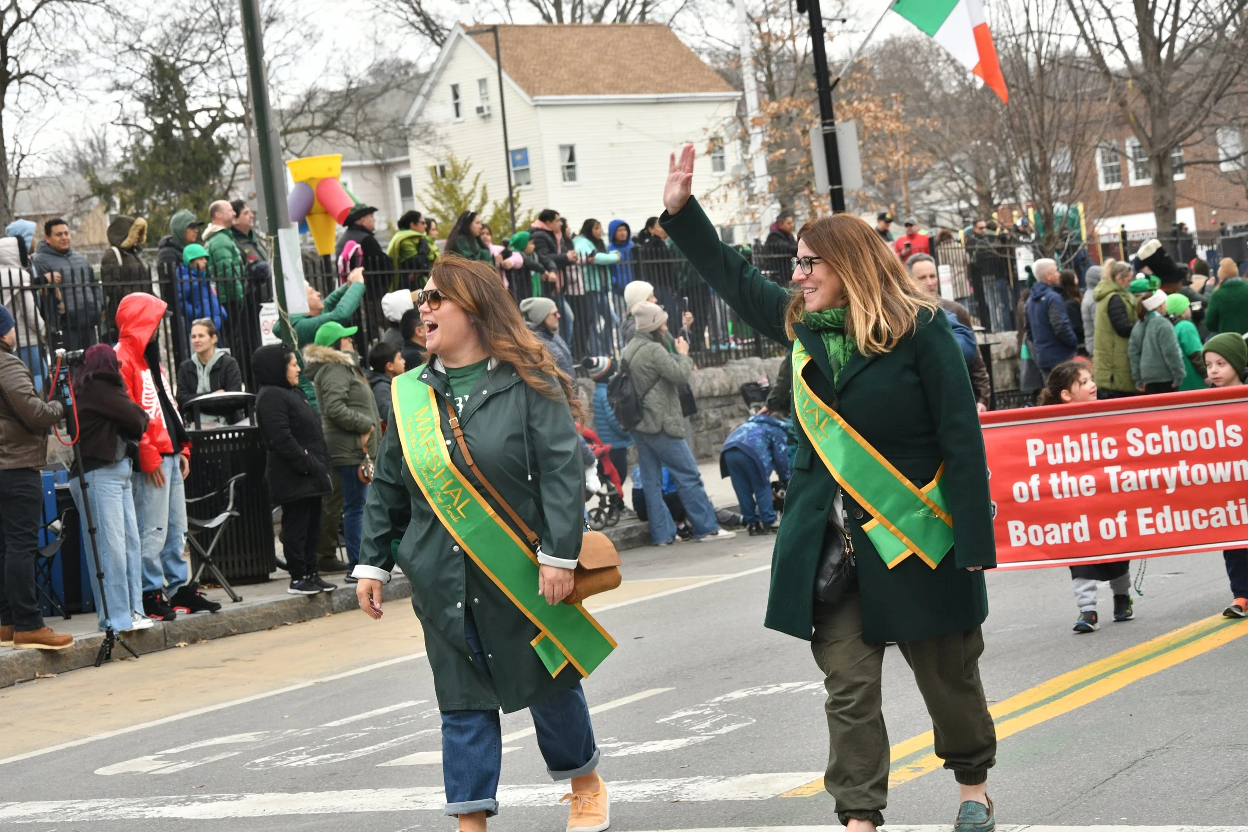 Two women walking in a parade, wearing green jackets and sashes, one waving to the crowd, with a crowd of people behind a fence and houses in the background.