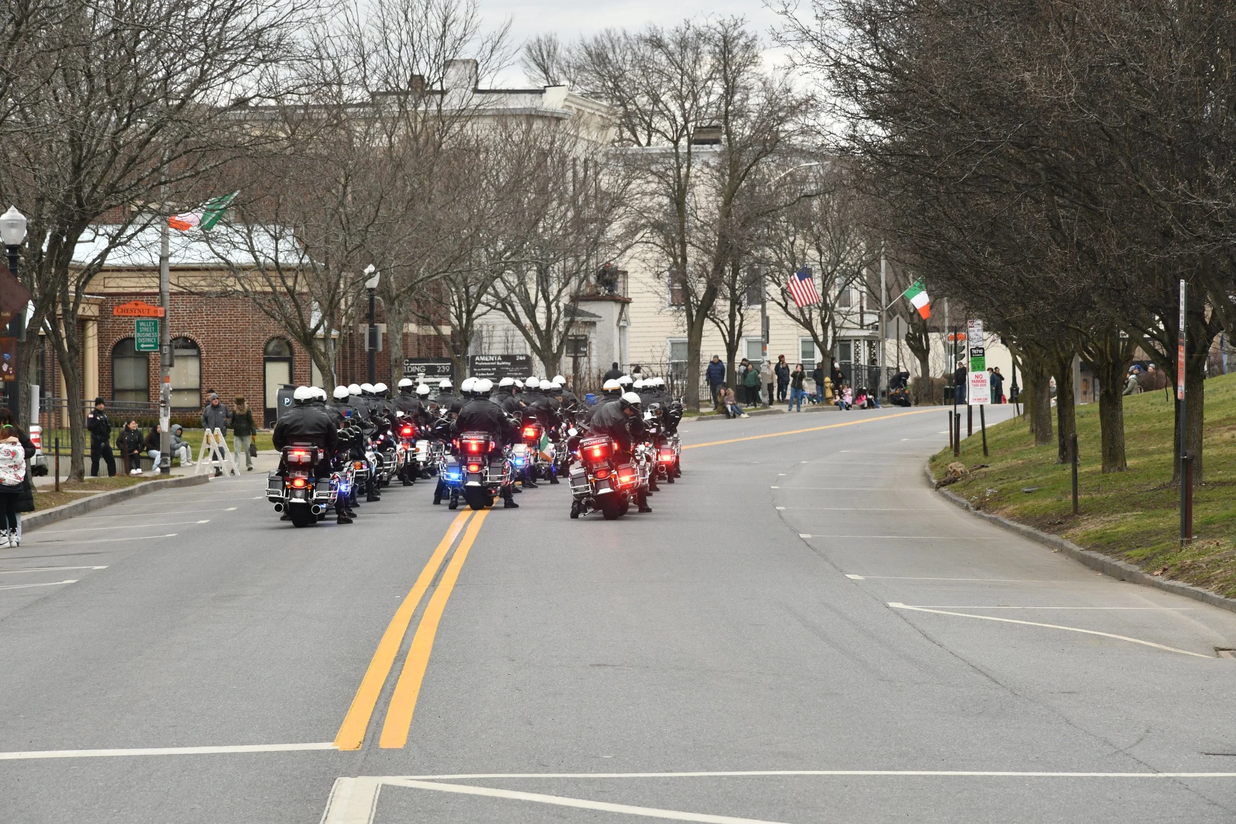 A group of police motorcycles with flashing lights driving down a street during a public event, with spectators lining the sidewalk and houses decorated with flags in the background.