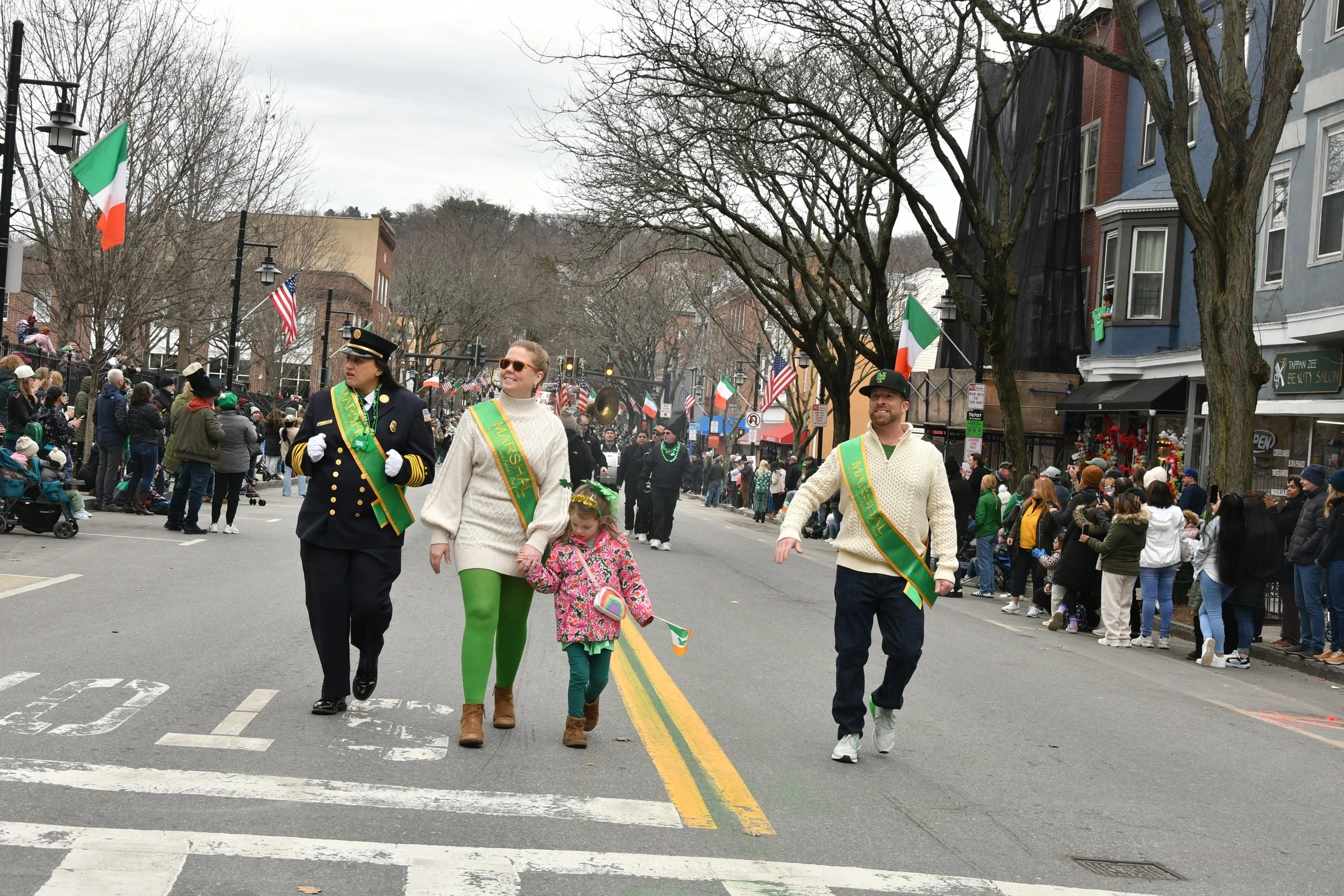 People participating in a St. Patrick's Day parade, some with Irish flags, wearing green, walking on a street with spectators on the sidewalk, leafless trees, and buildings in the background.
