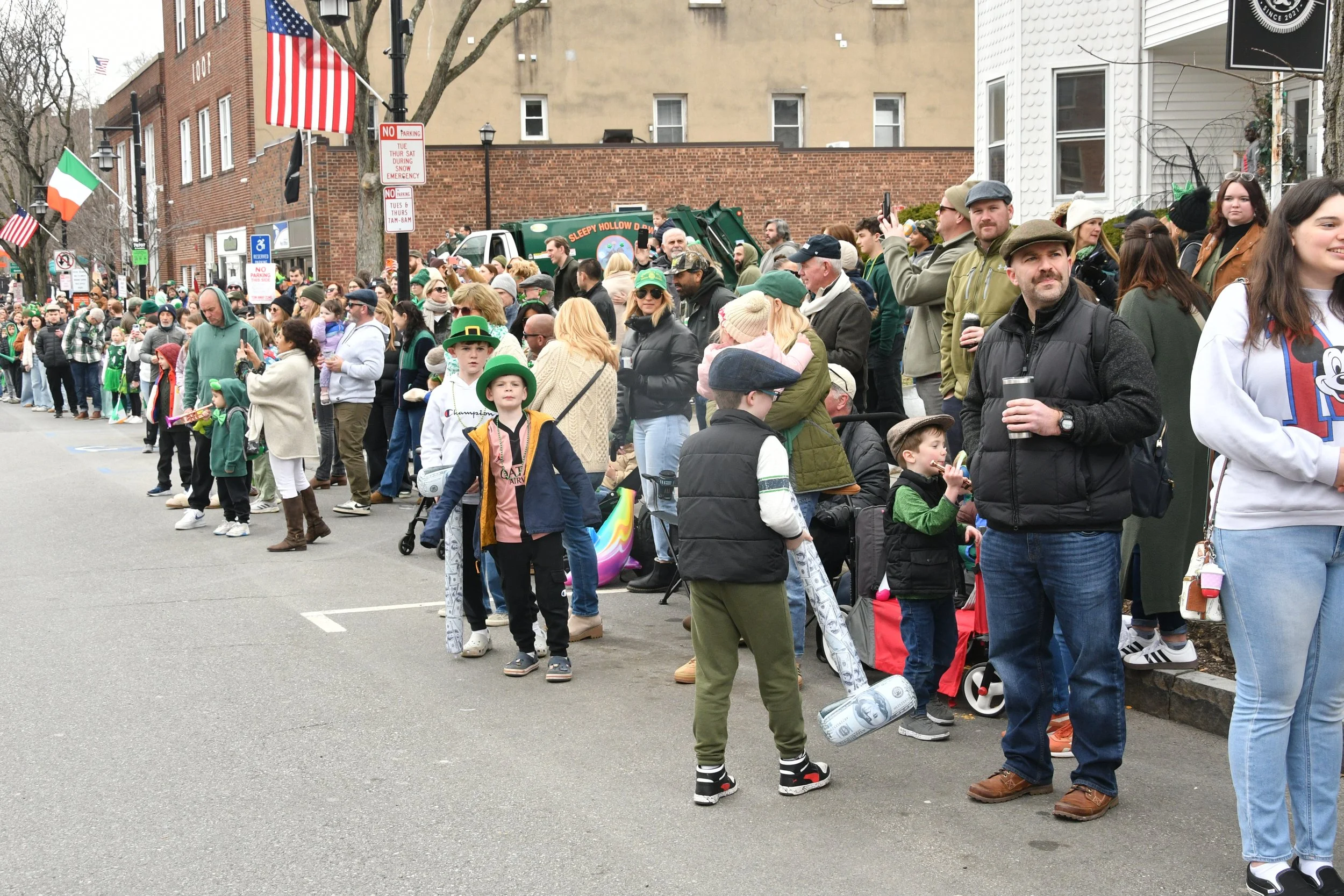 A large crowd of diverse people gathered outdoors, many dressed in green, celebrating St. Patrick's Day. Some are wearing hats, including leprechaun hats, and festive attire. There are flags, including American and Irish flags, and a garbage truck in