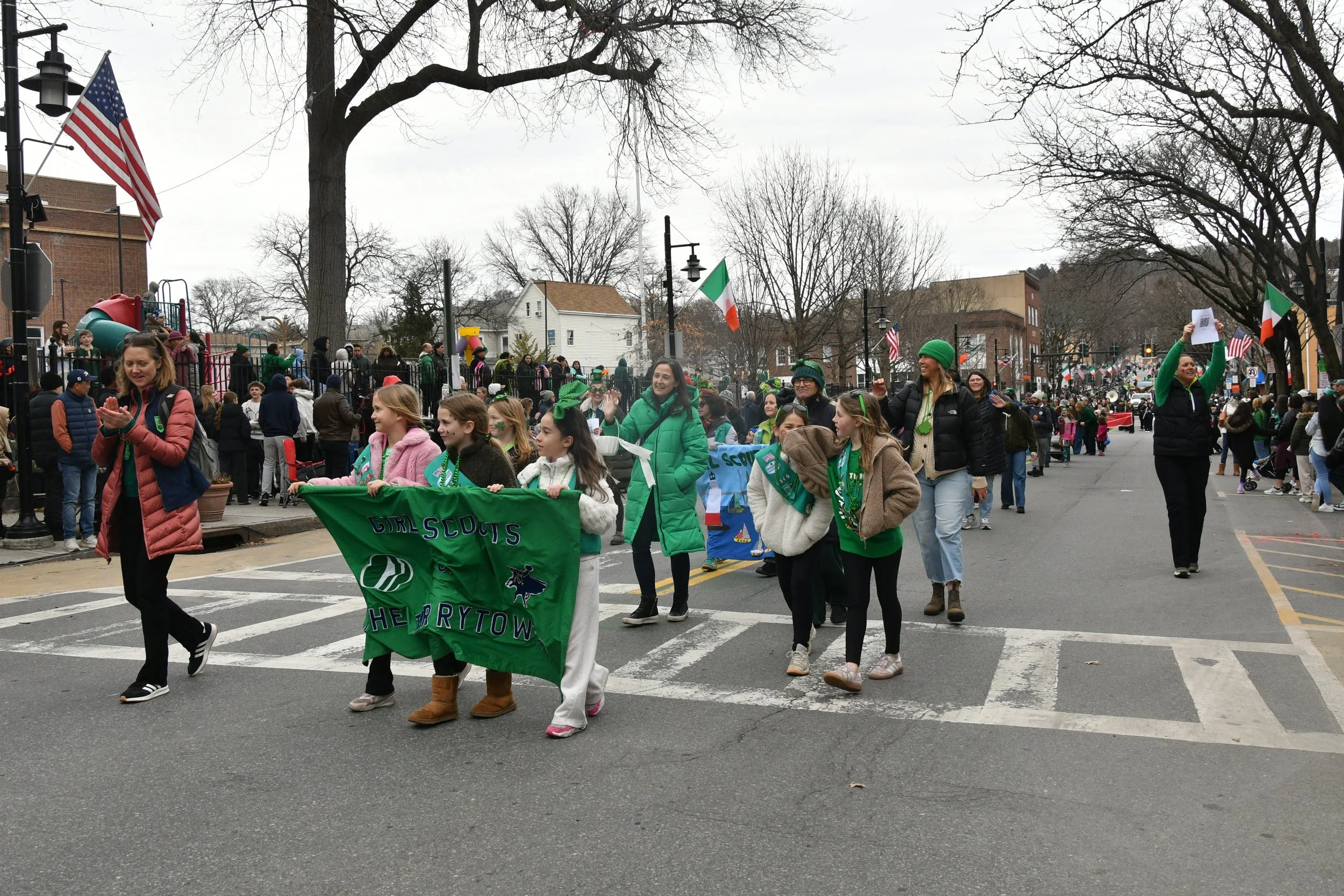 A parade with people walking down a street in a town, many wearing green and holding a green banner that reads "Girl Scouts" and "The Berytown." The parade has American and Italian flags, and people are watching from the sidewalks and a playground ar