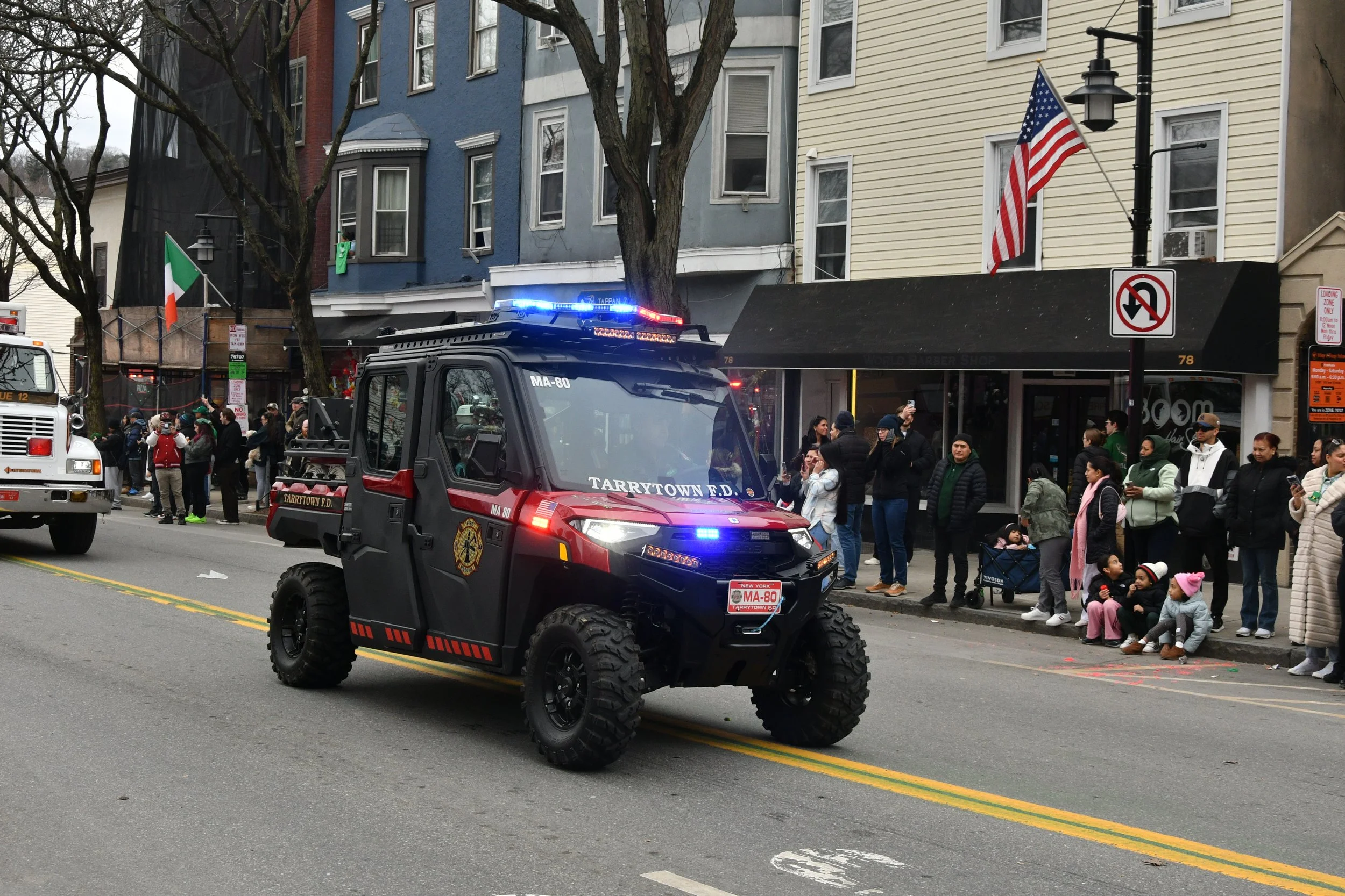 A parade with a black rescue vehicle labeled Tarrytown Fire Department in the foreground, and a crowd of people on the sidewalk watching. There are buildings with colorful facades behind the crowd, and flags flying, including the United States flag a