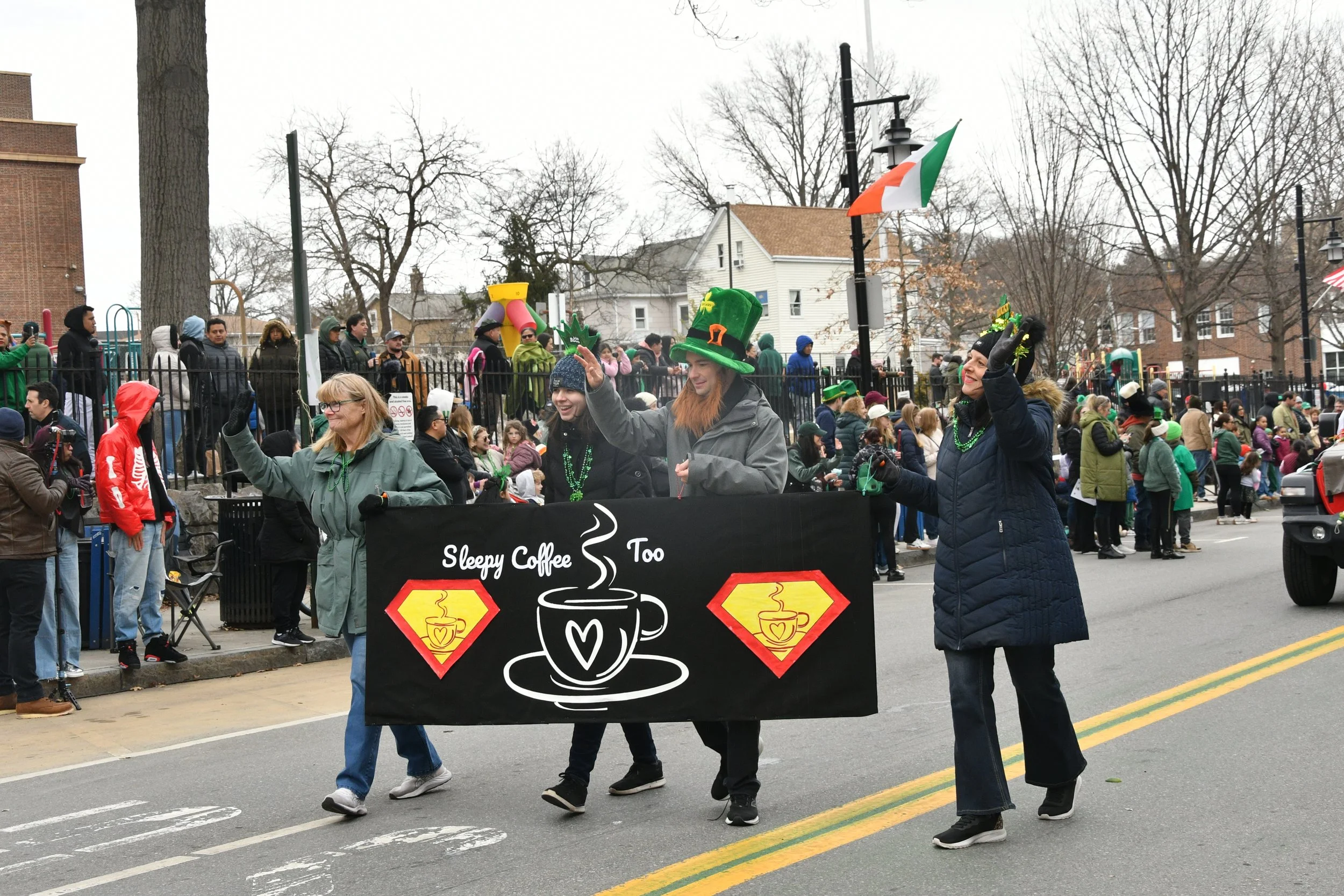 People participating in a parade, some wearing St. Patrick's Day accessories, passing by a Sleepy Coffee tent on a city street. The street is lined with leafless trees and buildings.