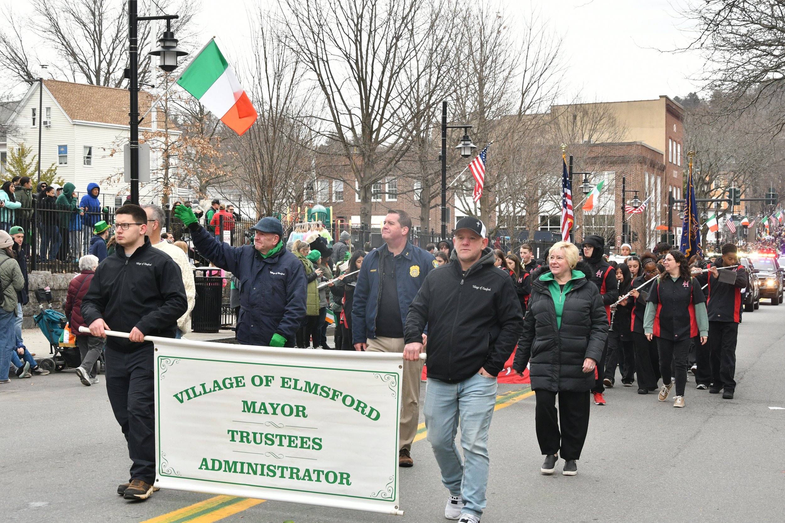 People participating in a parade or march, holding flags including the Irish flag and American flags, with some wearing green and other colors, in a small town setting with trees and buildings in the background.
