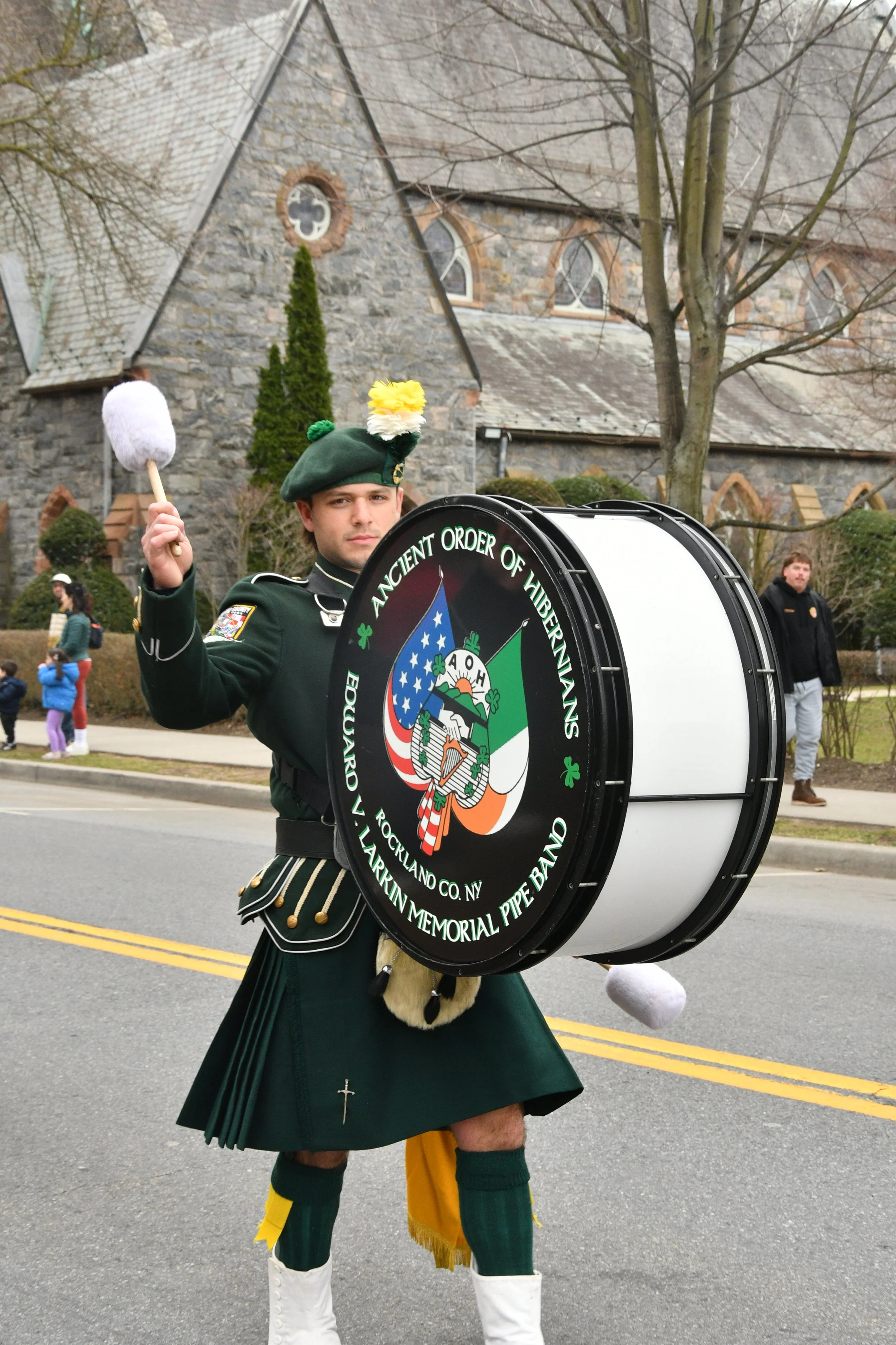 A man dressed as a traditional Scottish bagpiper, wearing a dark green kilt, a green sporran, and white knee-high boots, playing a large drum during a parade or procession on a street. In the background, there is a stone church with arched windows an