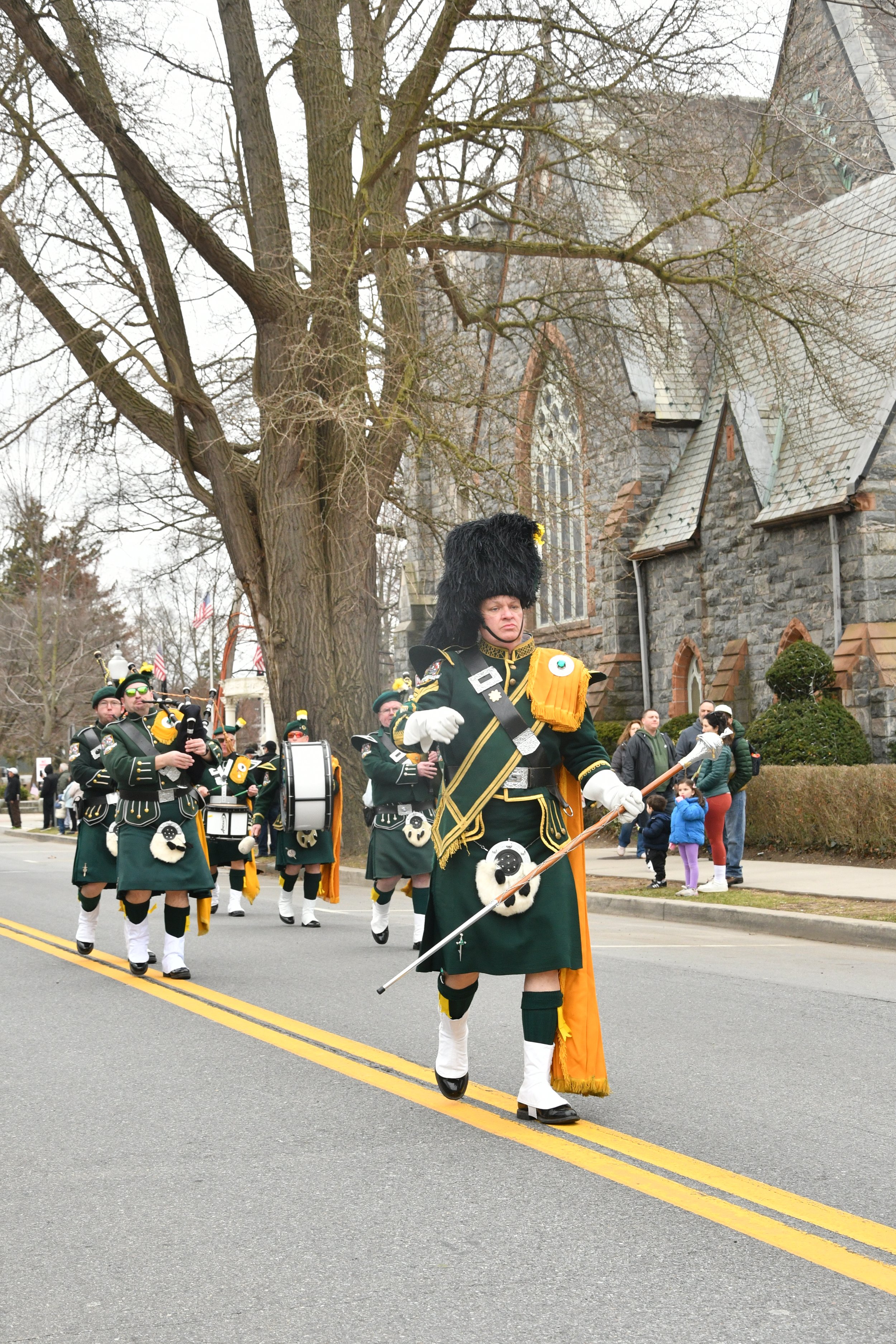 A marching band dressed in green and orange uniforms, playing instruments, part of a parade on a city street in front of a stone church with many spectators, including children.