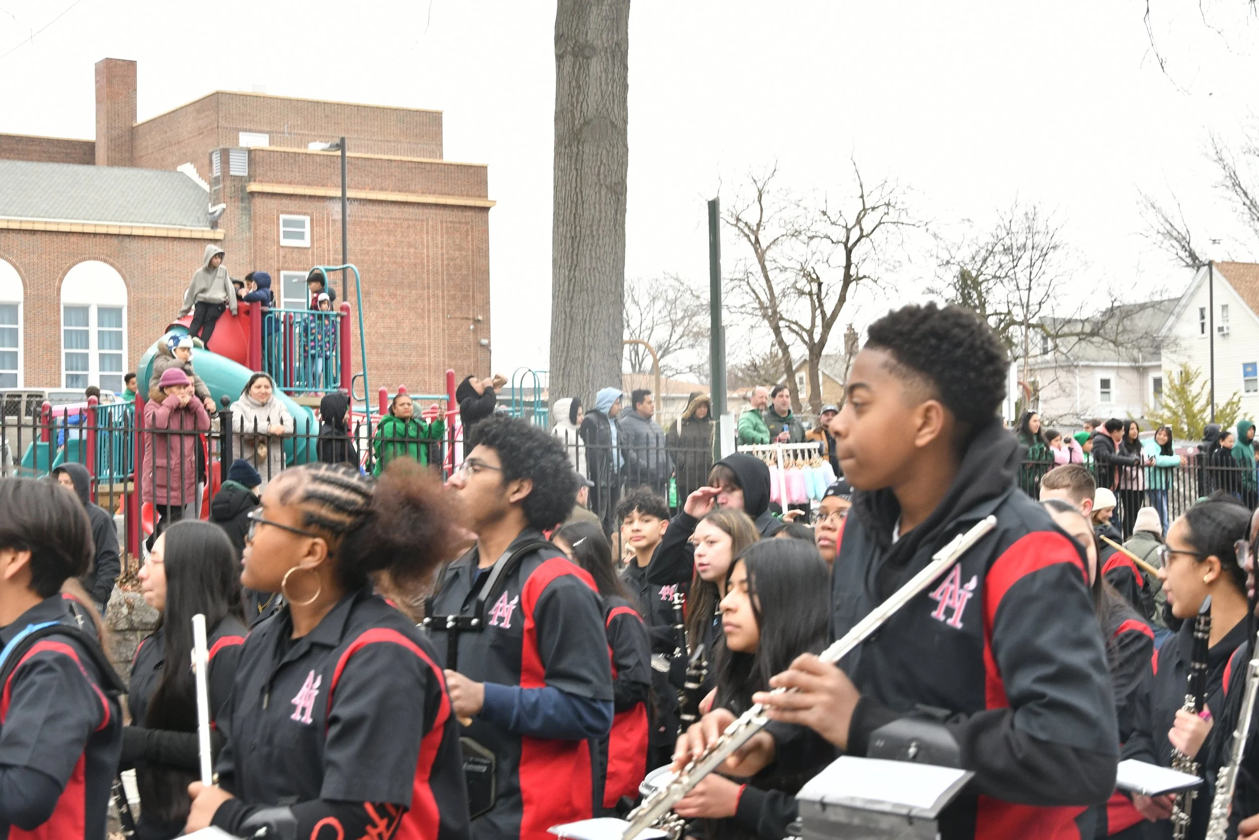 A large group of people gathered outside in a public space, some walking and others watching a parade or event. In the background, children are playing on a playground set with a slide and climbing structure, surrounded by brick buildings and leafles