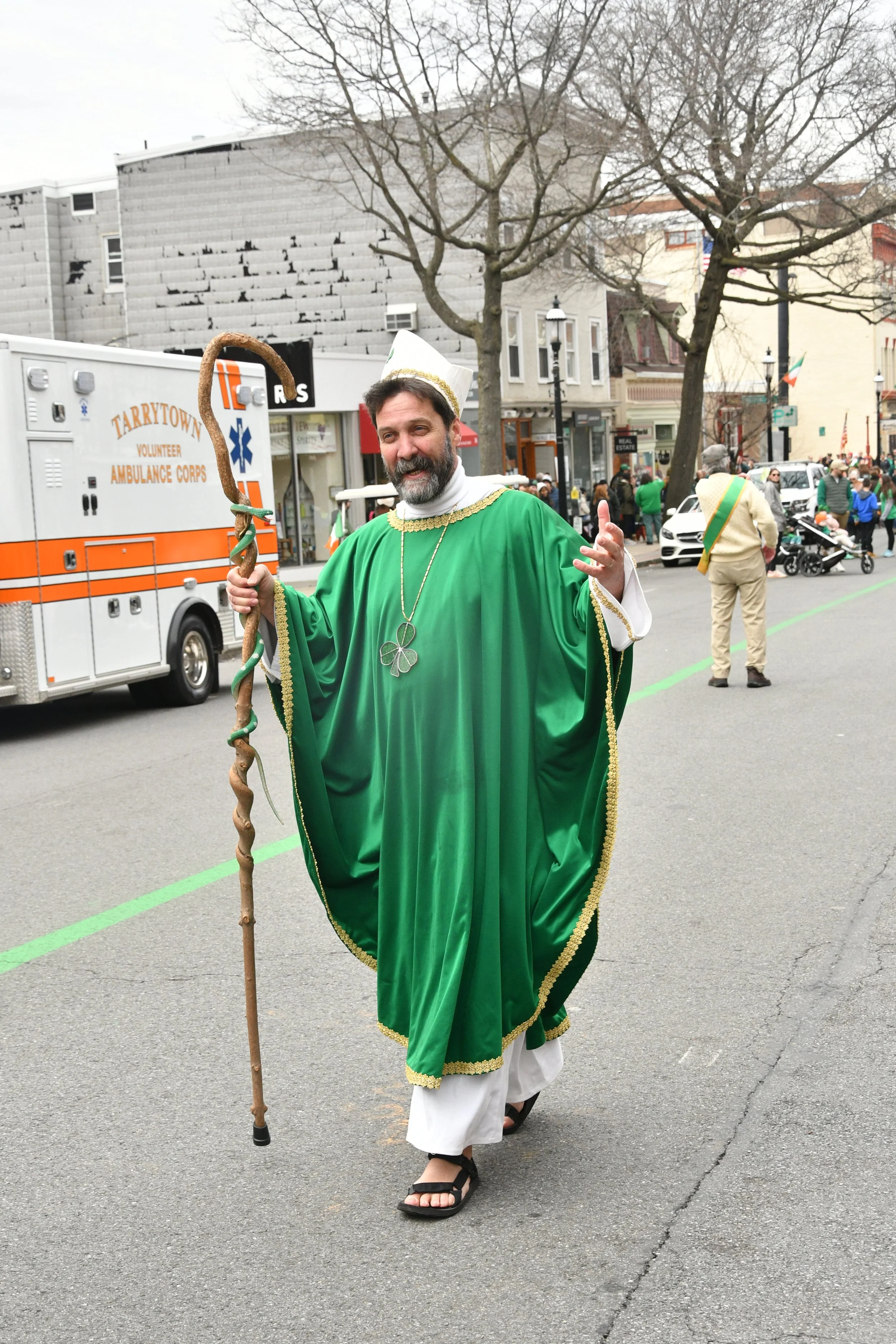 Man dressed as St. Patrick in a green robe with gold trim, holding a wooden staff, participating in a parade on a city street, with an ambulance and onlookers in the background.