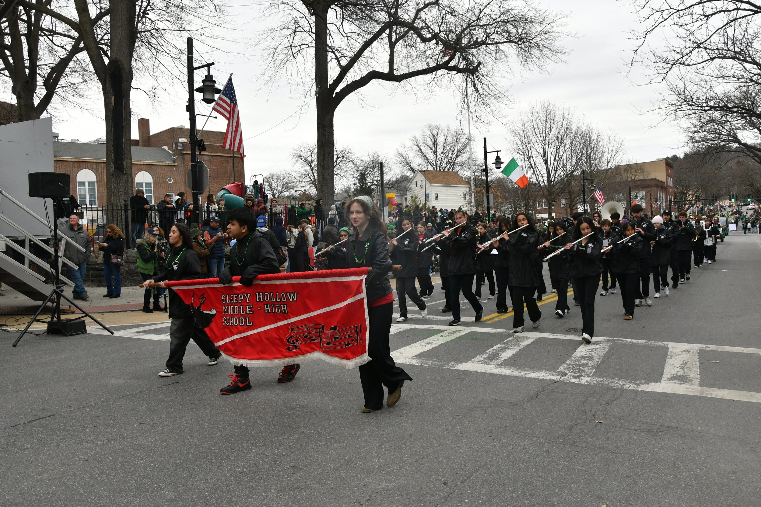 A parade with students from Sleepy Hollow Middle and High School marching with musical instruments, including flutes. The students wear black clothing, and some wear beads. An American flag and an Irish flag are displayed, and onlookers are watching 