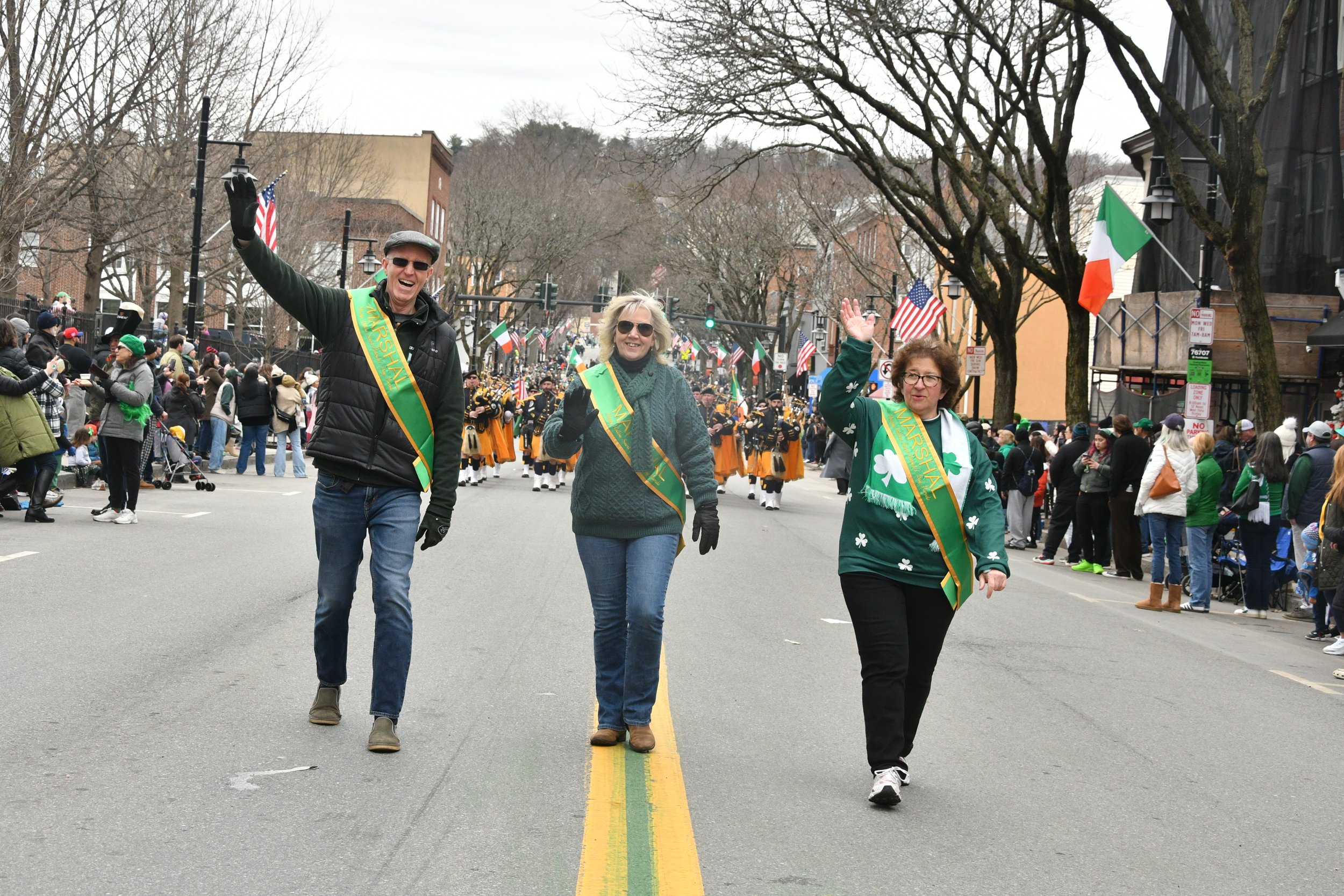 Three people walking on a city street during a parade, waving and smiling, with a marching band in the background and wrapped buildings and trees lining the street.