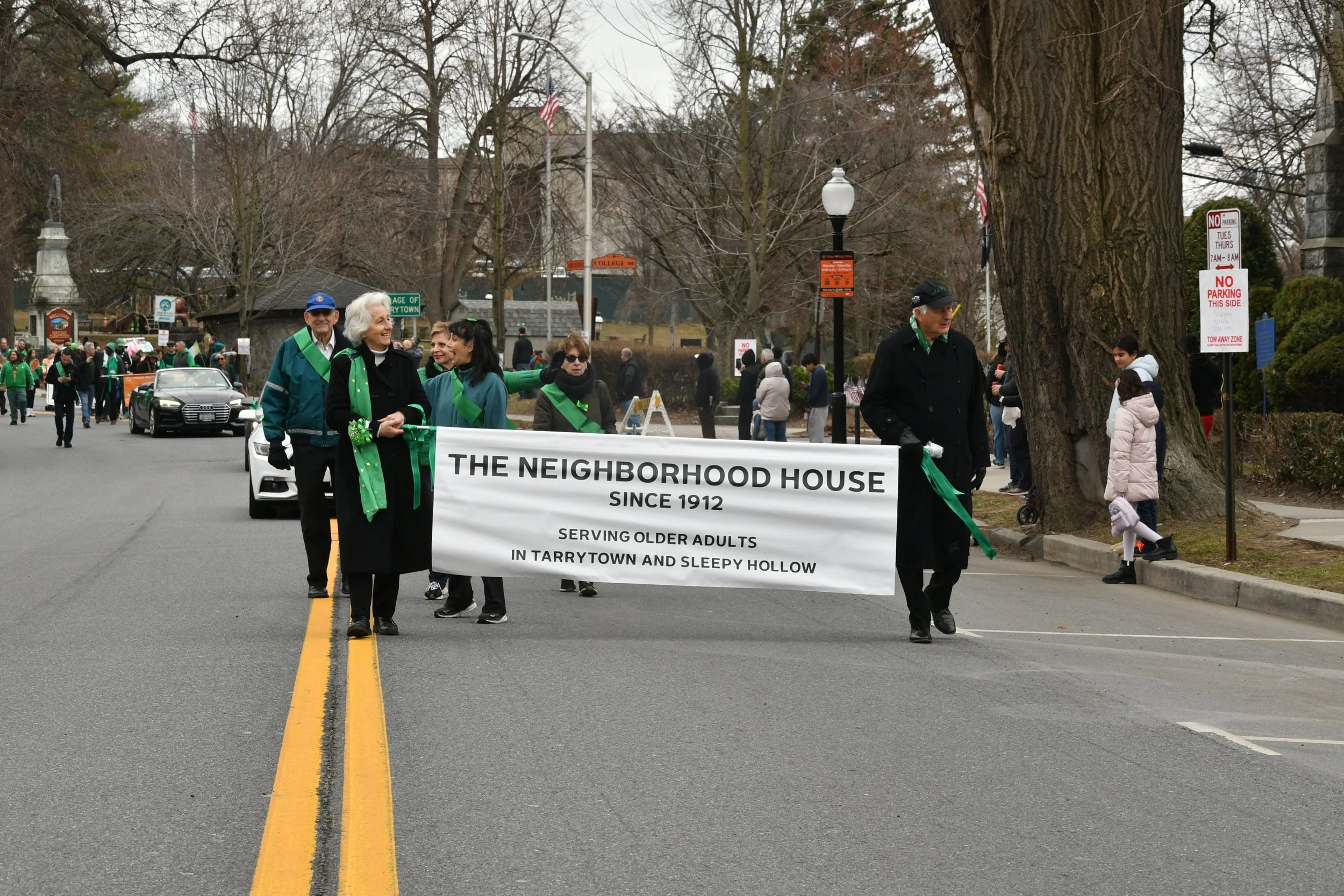 A parade march with people holding a banner that reads 'The Neighborhood House since 1912, serving older adults in Tarrytown and Sleepy Hollow', moving down a street lined with Trees and bystanders.