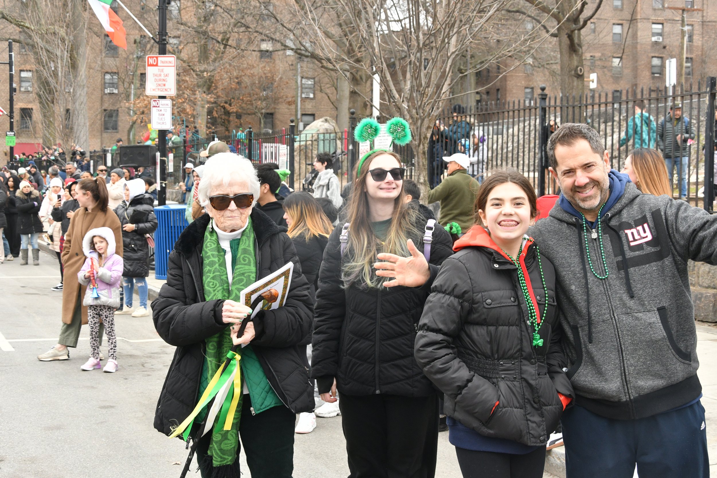 A diverse group of people celebrating outdoors during a festive event, with some wearing green accessories and St. Patrick's Day attire. In the foreground, a smiling young girl and an older woman holding a program, with others in the background parti