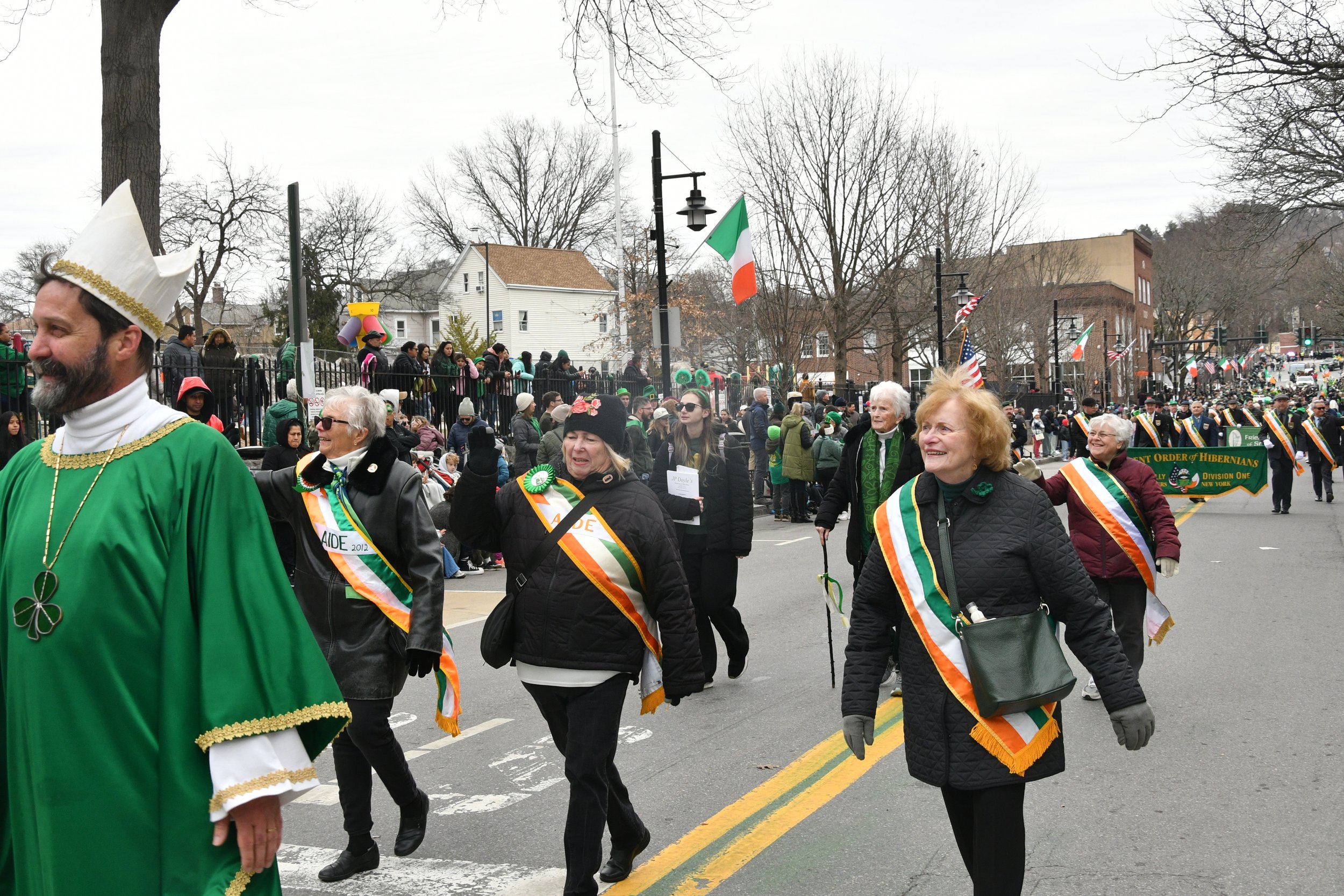 People participating in a St. Patrick's Day parade, dressed in festive attire including green clothing, hats, and sashes, walking down a city street lined with leafless trees and a crowd of spectators.