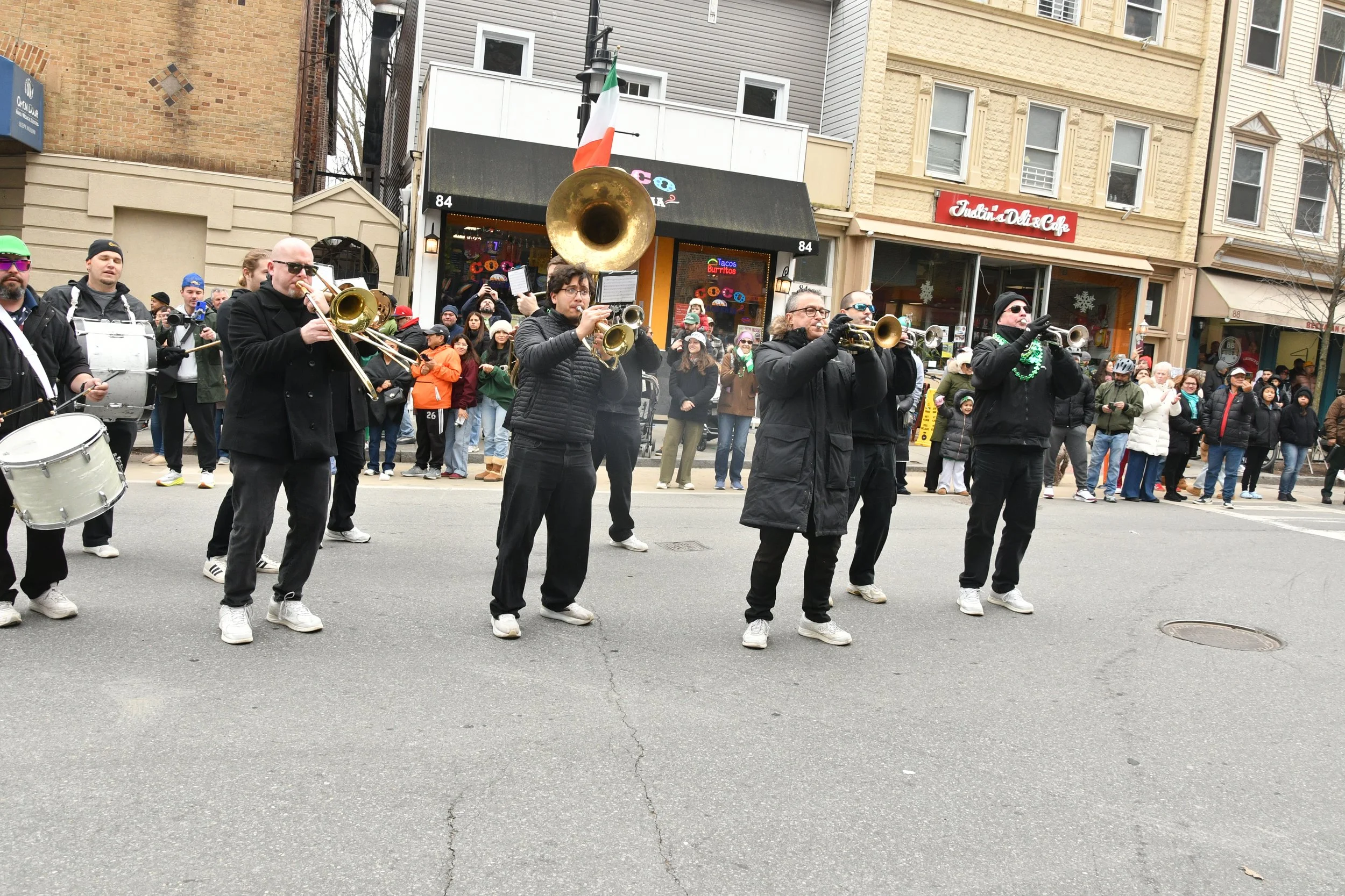 A marching band performs on a city street with an audience watching. The band members are playing wind instruments and drums, wearing black clothing. Behind them are various storefronts, including a cafe and a deli.