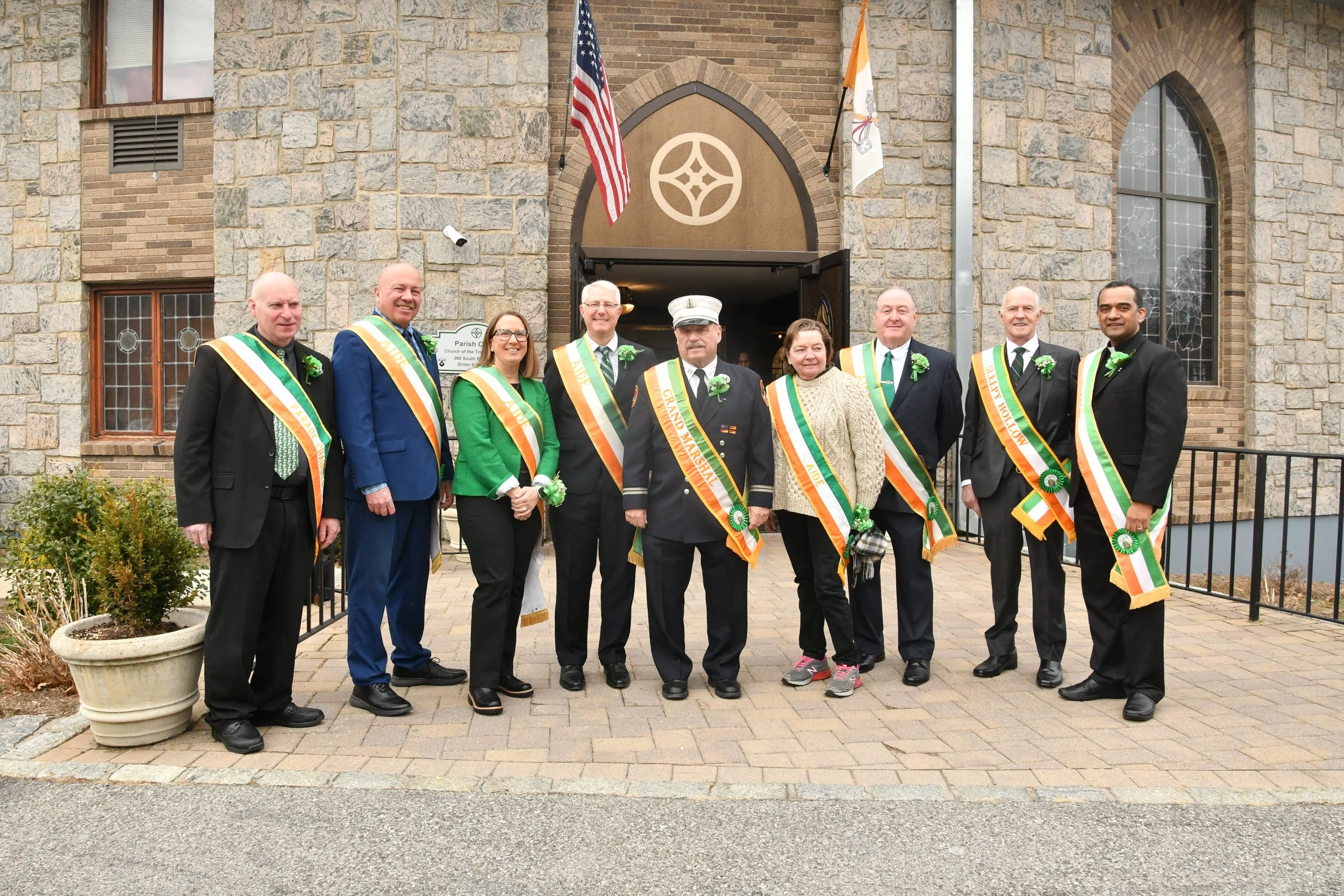 A group of nine people standing in front of a church entrance, wearing formal attire with sashes and flowers, inside a stone building with flags and windows.