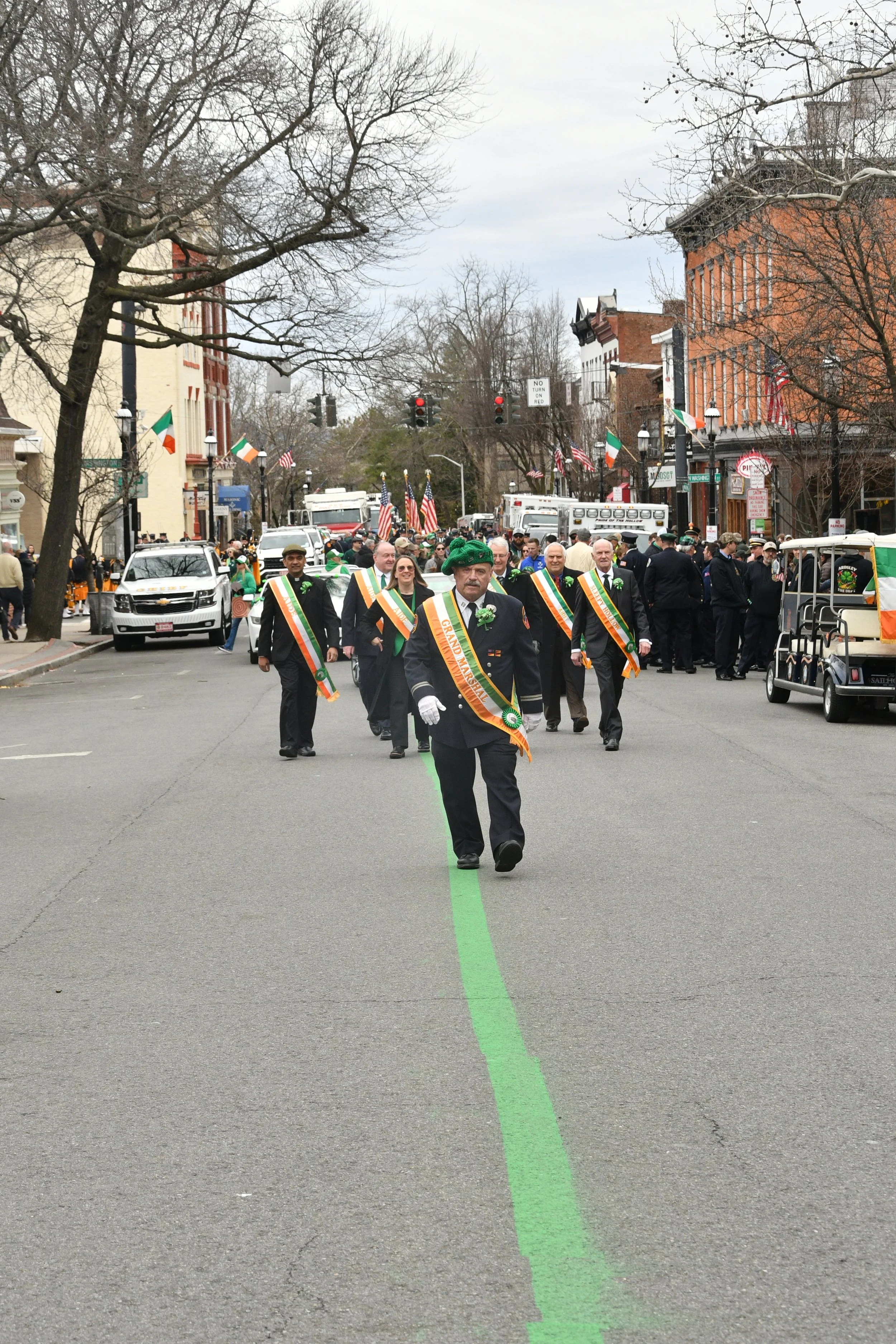 People participating in a parade or march on a city street decorated with small flags, with some individuals wearing sashes and uniforms, and a large green line down the center of the road.