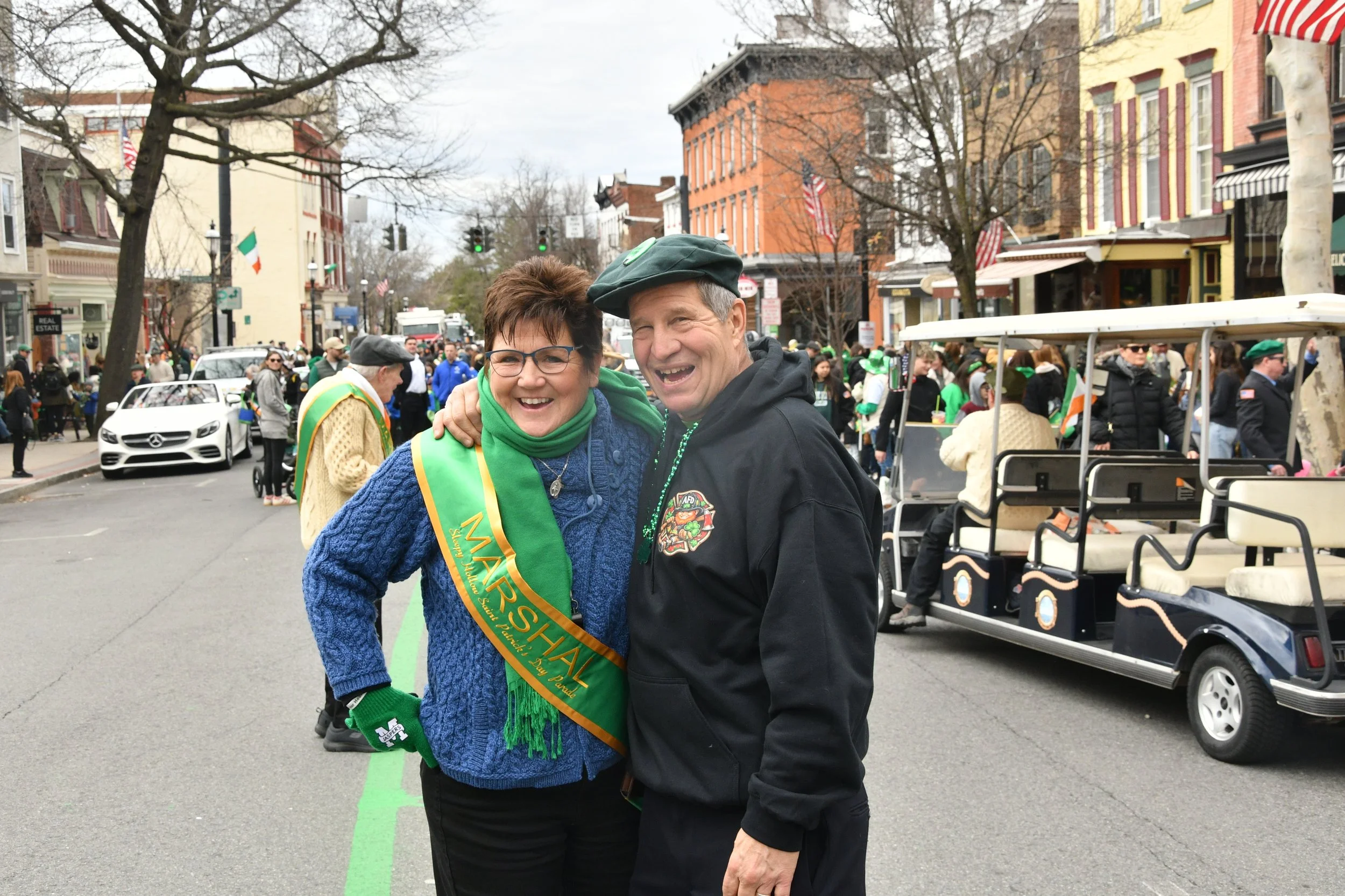 Two people smiling and greeting each other during a parade or festive event on a city street. The woman on the left is wearing glasses, a blue sweater, and a green sash that says 'MARCH'. The man on the right is wearing a black jacket and a green ber