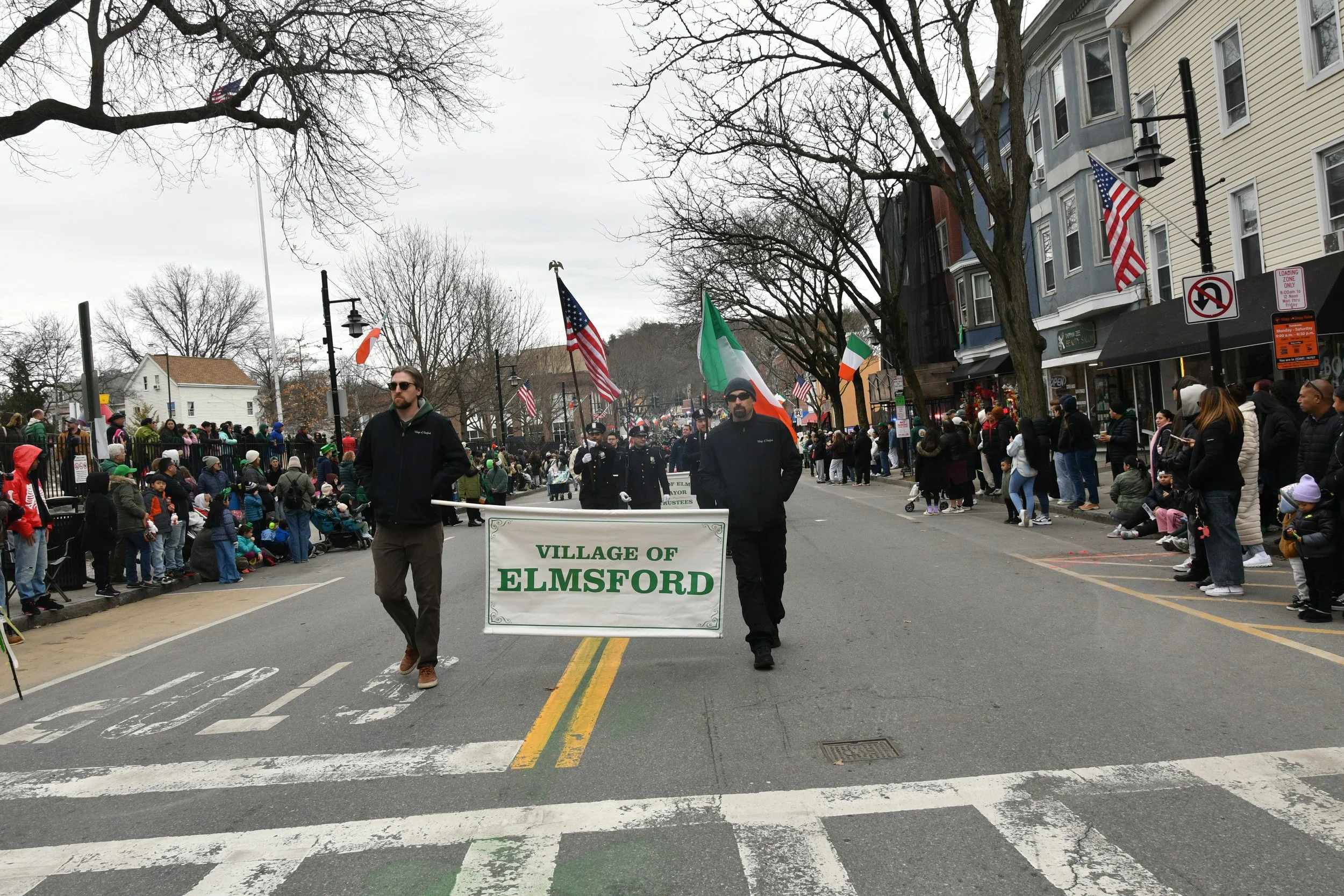 A parade with people walking down a street holding a sign that reads 'VILLAGE OF ELMSFORD'. There are many spectators on both sides of the street, some sitting and some standing. Flags, including American and Irish flags, are visible. The weather is 