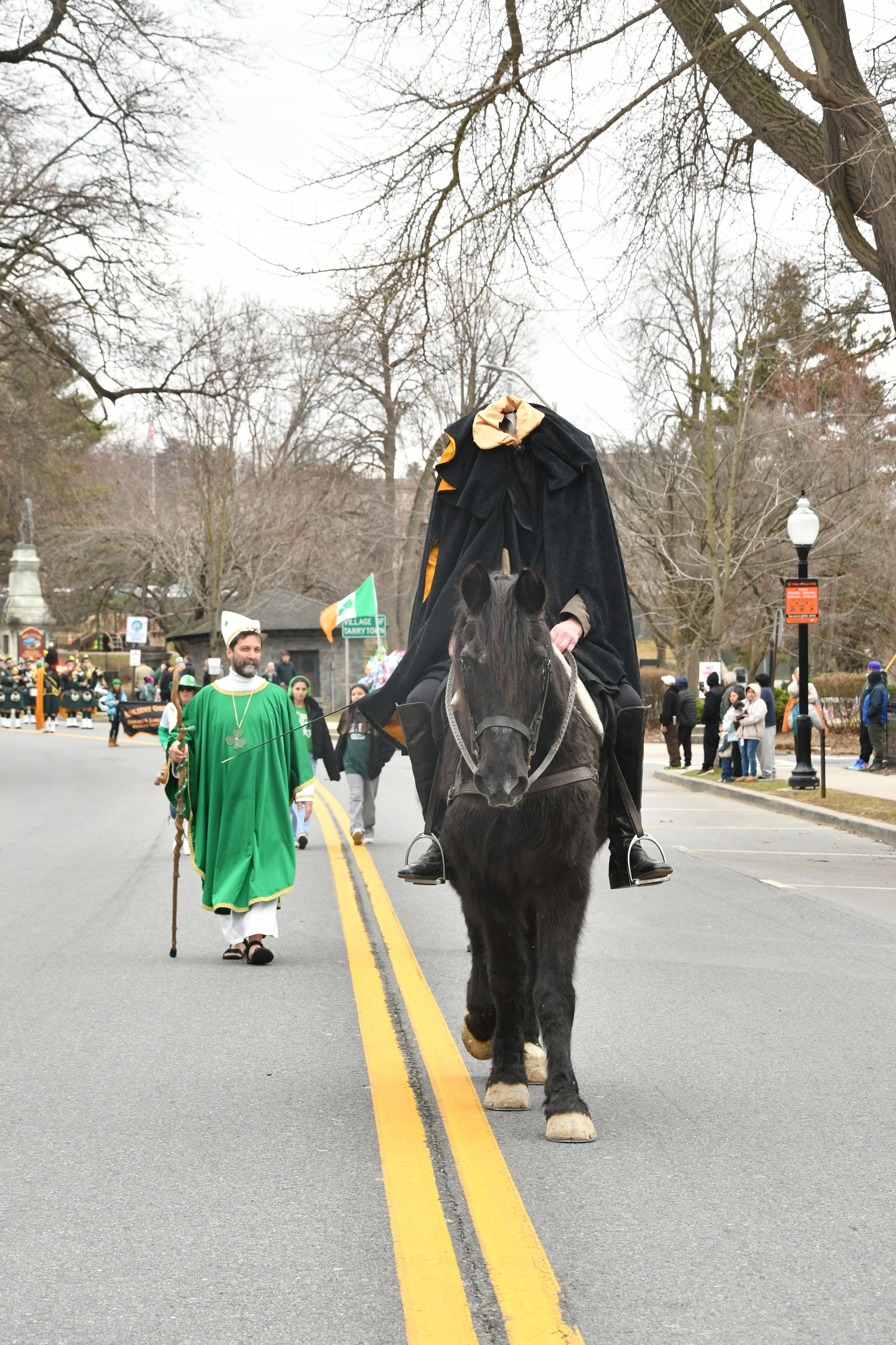 People participating in a parade, including a man riding a horse, dressed in black, and wearing a hat, with other spectators and parade participants visible in the background, overcast sky, leafless trees.