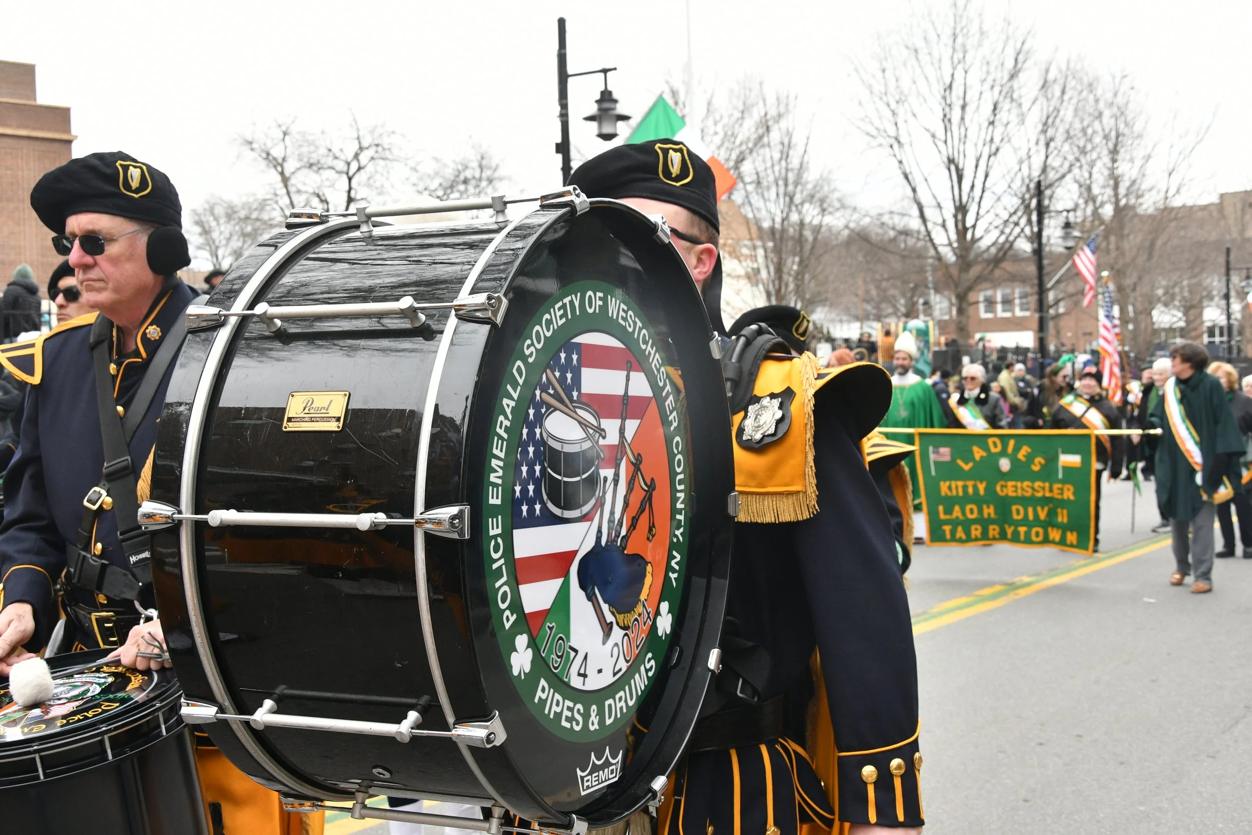 Street parade with musicians and spectators, person in uniform playing drums, American flags and parade banners visible.
