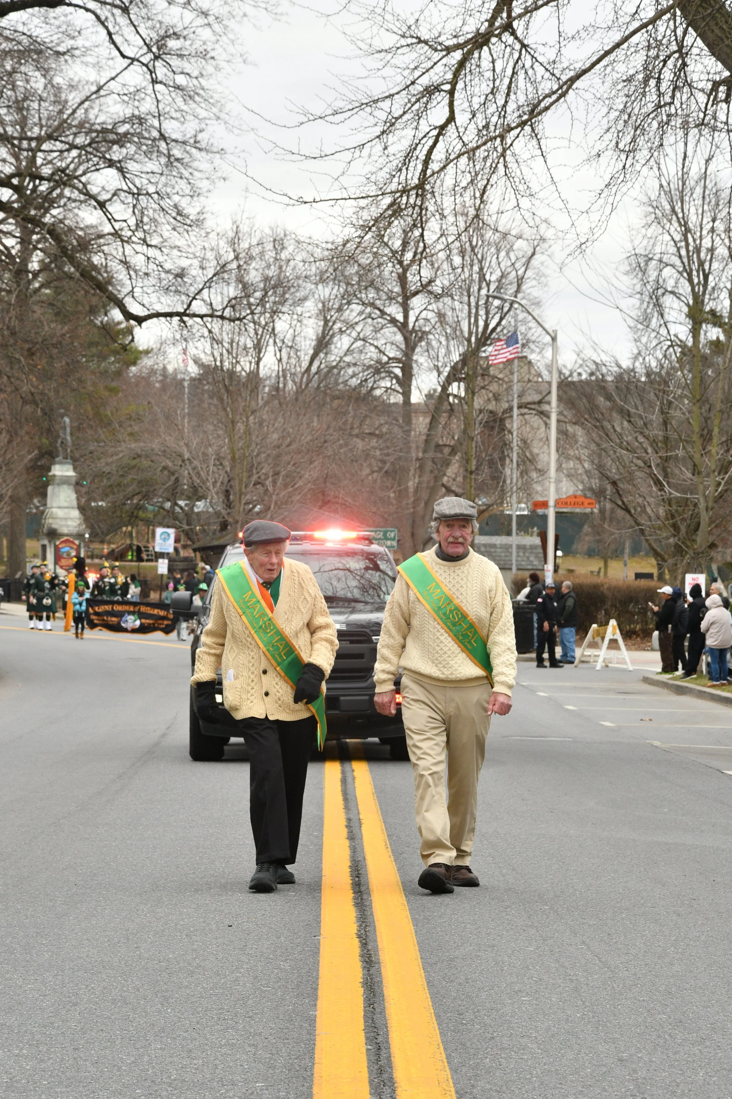 Two elderly men wearing cream-colored sweaters and marshals sashes walk down the street during a parade or event, with a background of leafless trees, onlookers, a police car, and American flags.