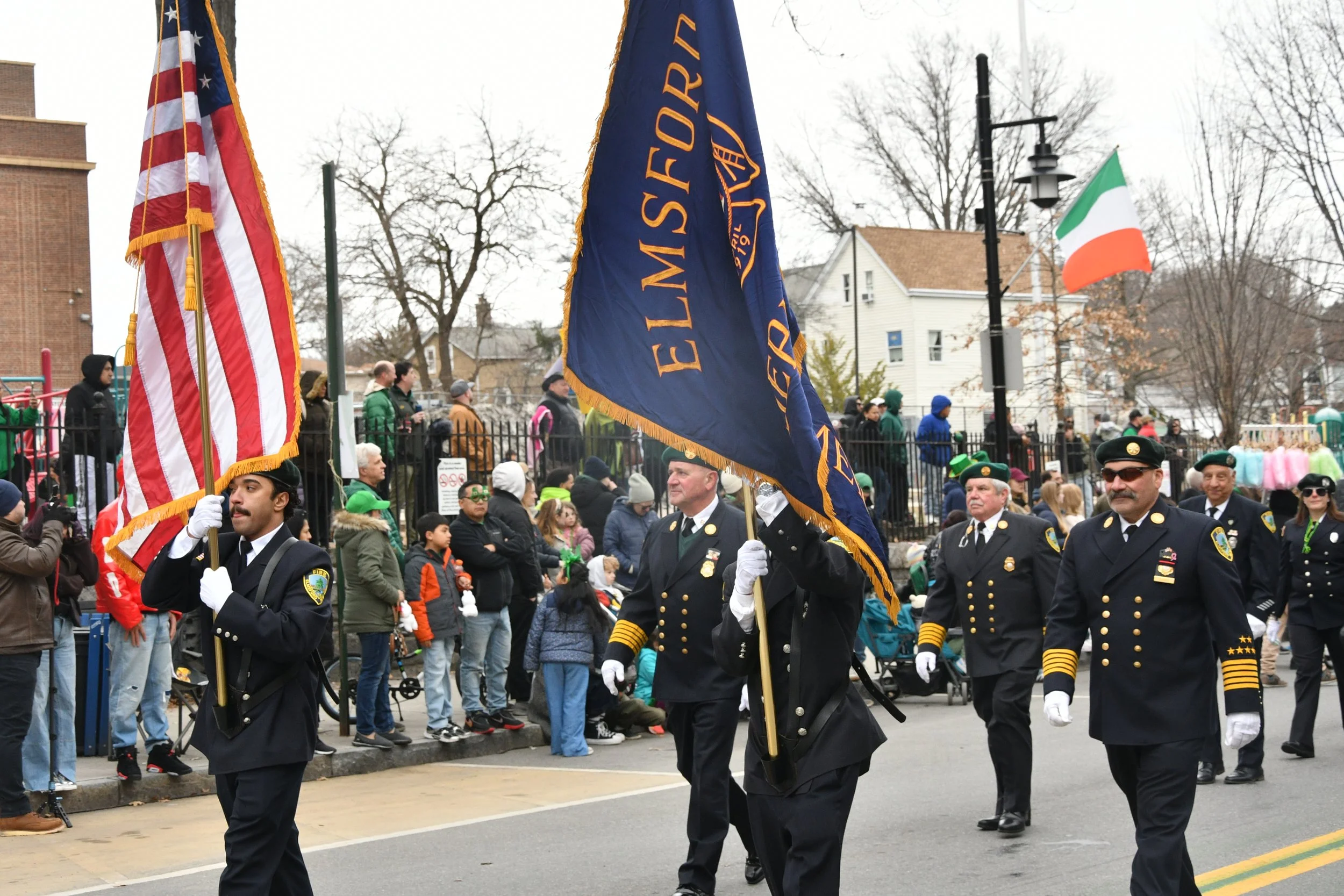 Marching parade with uniformed individuals carrying flags, including an American flag and an Irish flag, during a public event with spectators and onlookers.