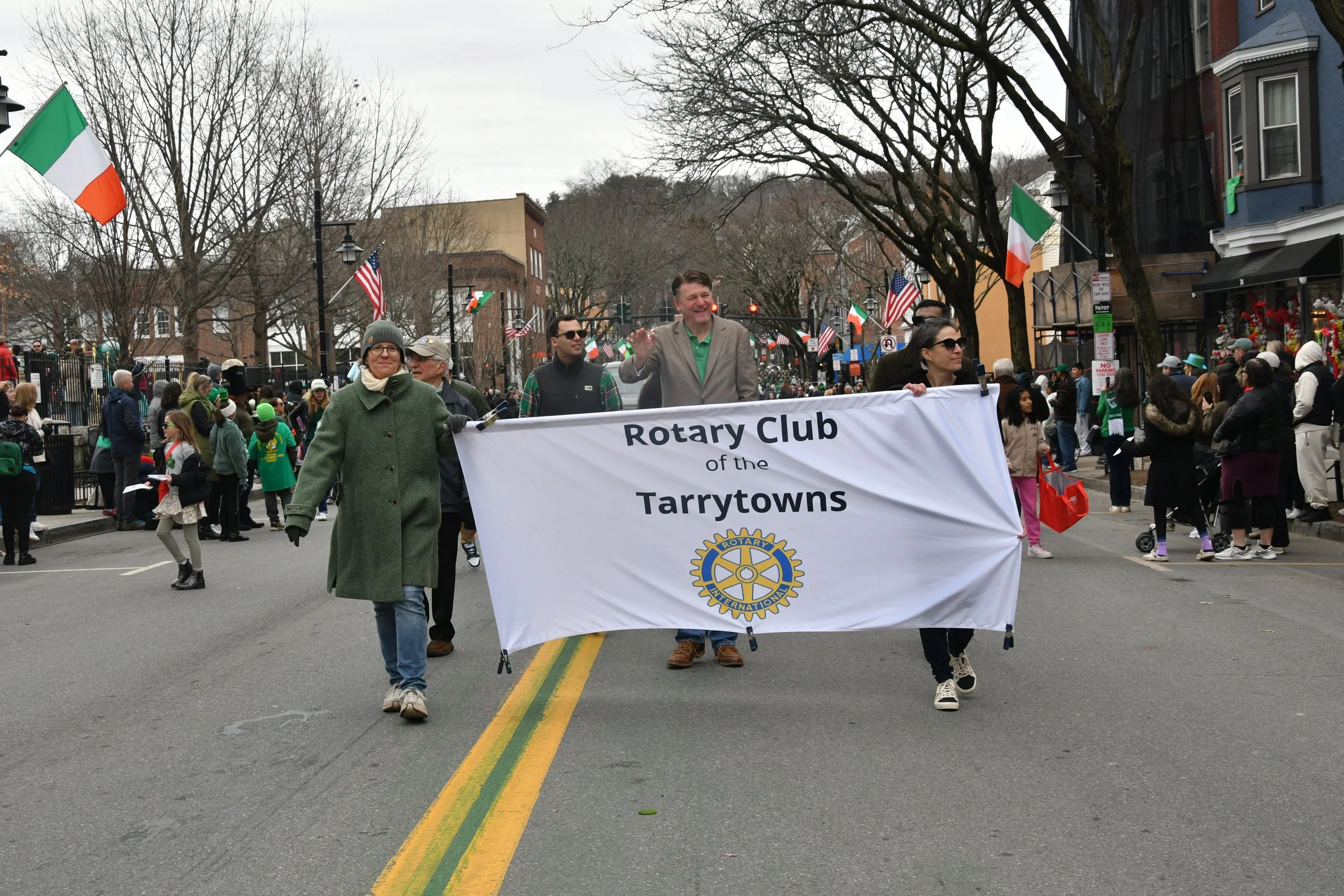 A parade on a city street with people holding a banner that reads 'Rotary Club of the Tarrytowns' and flags of the United States and Ireland, with spectators watching along the sidewalk and decorated storefronts.