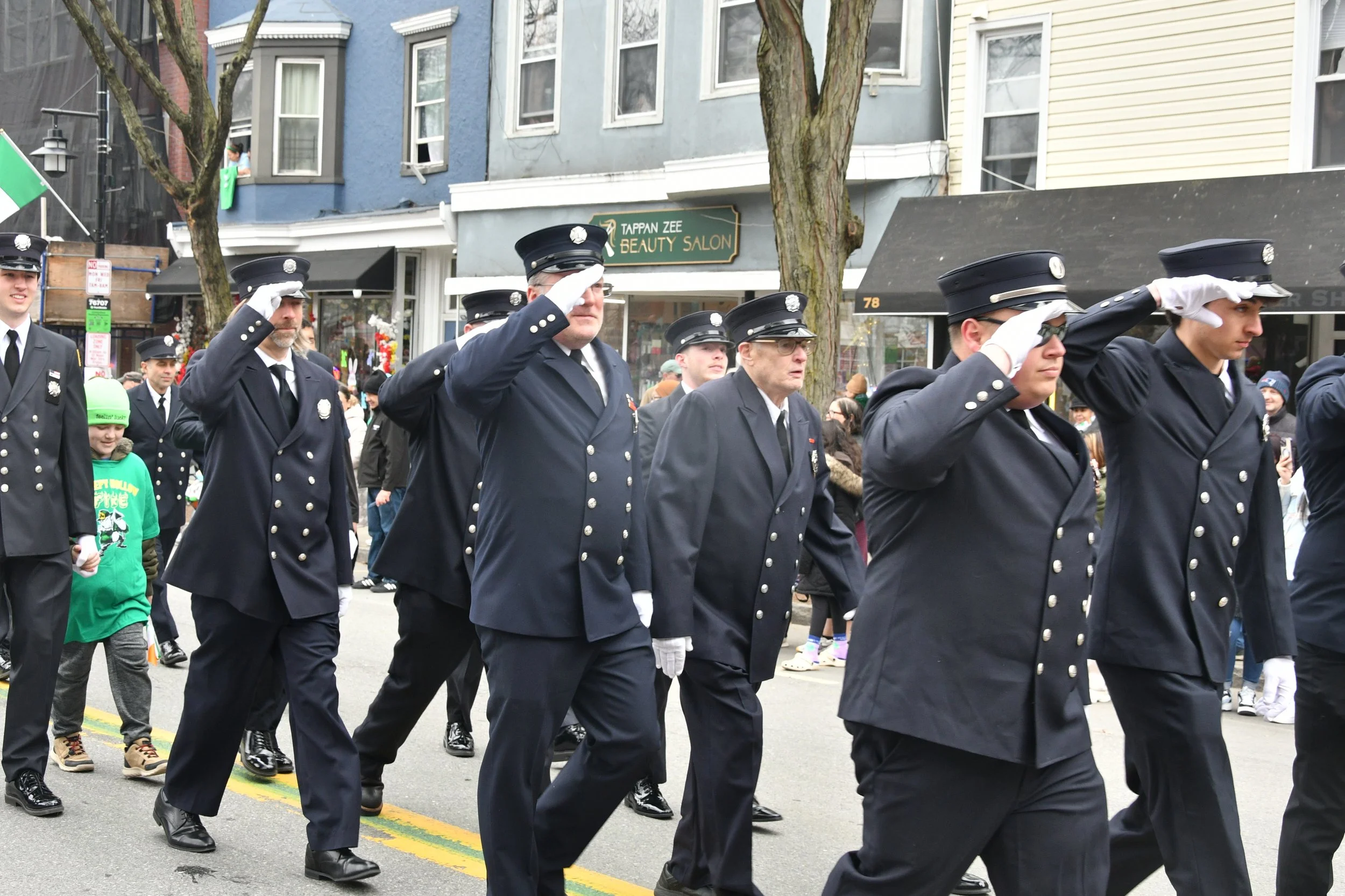 Group of uniformed police officers marching in a parade, saluting as they walk down a city street lined with buildings and trees.