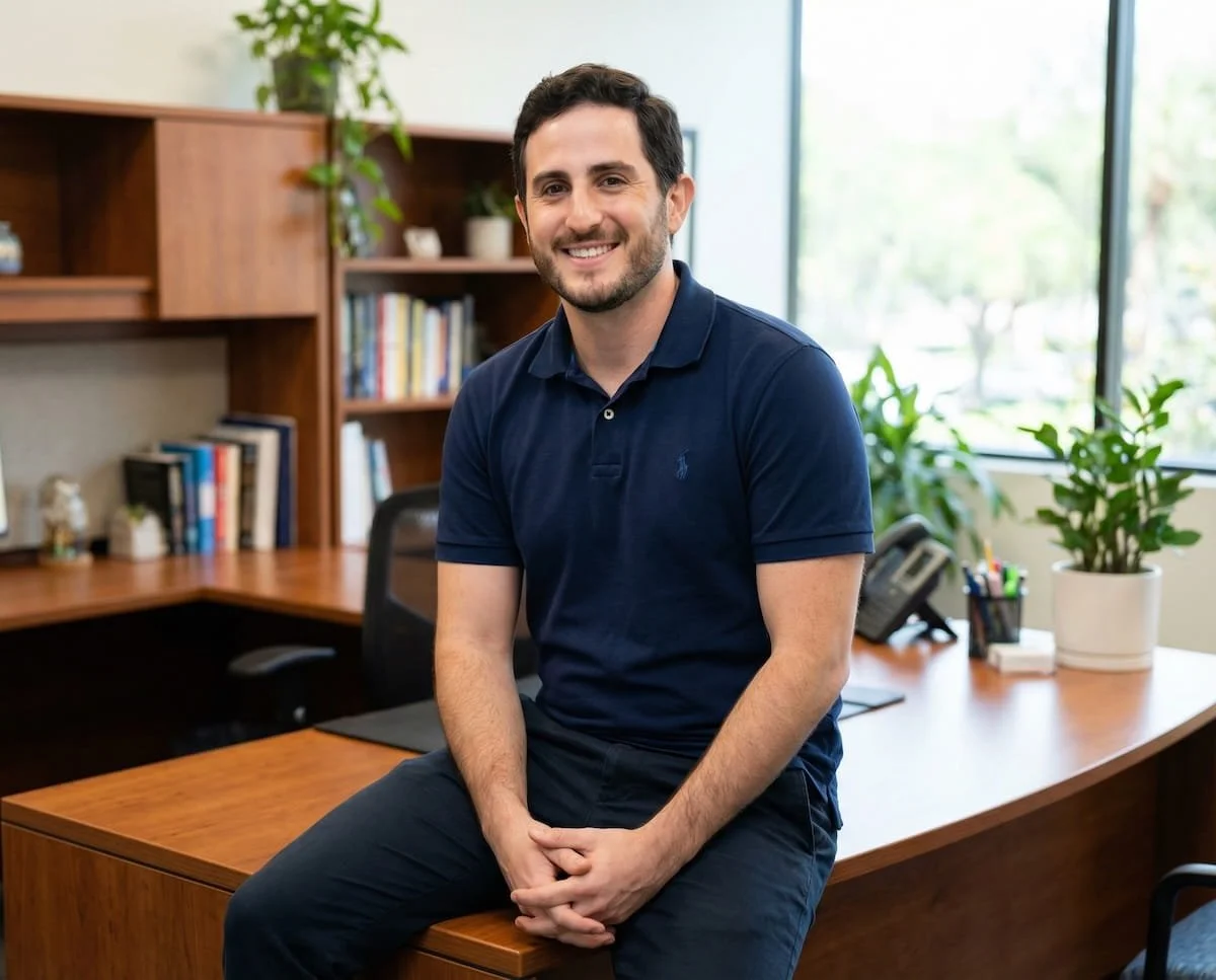 David Feingold, employment attorney and owner of DZ Law, in a navy blue polo shirt sitting on the edge of a wooden desk in a bright office with bookshelves, plants, and large windows.