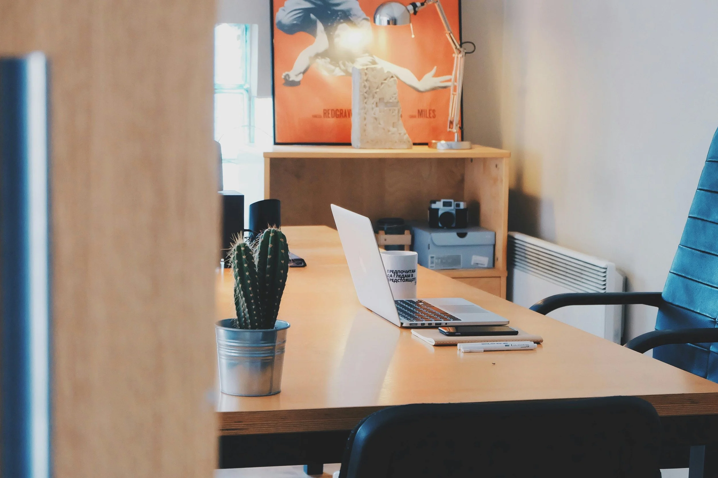An office workspace with a wooden desk, a cactus plant in a metal pot, a silver laptop, a notebook, a pen, and a white mug. In the background, a wooden shelf holds a camera and a plastic container, with a framed art piece of a running horse and a desk lamp on top. The room has beige walls, a chair with a blue cushion, and a radiator.