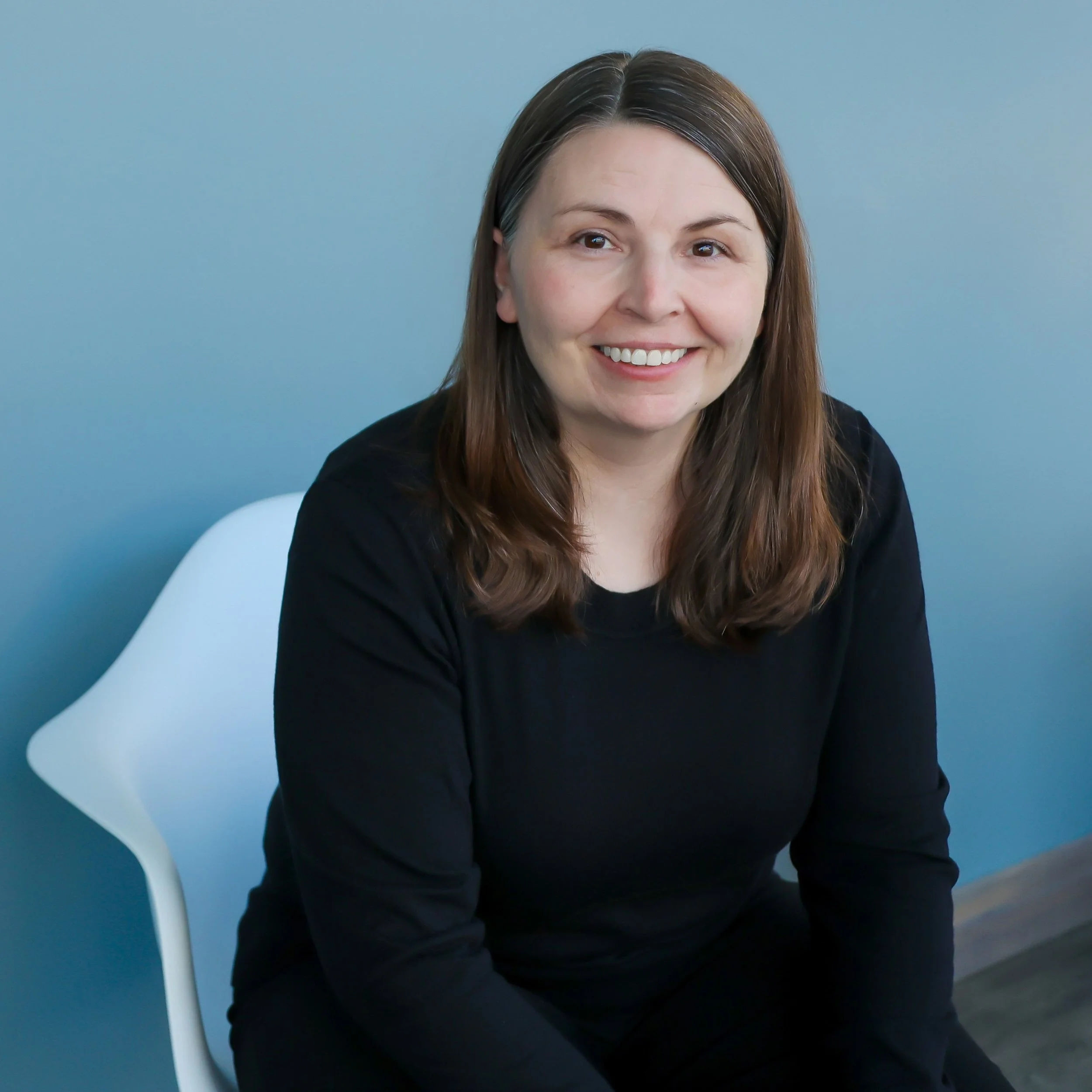 Photo of co-founder Alana Johnston sitting in white chair wearing black clothing against blue wall