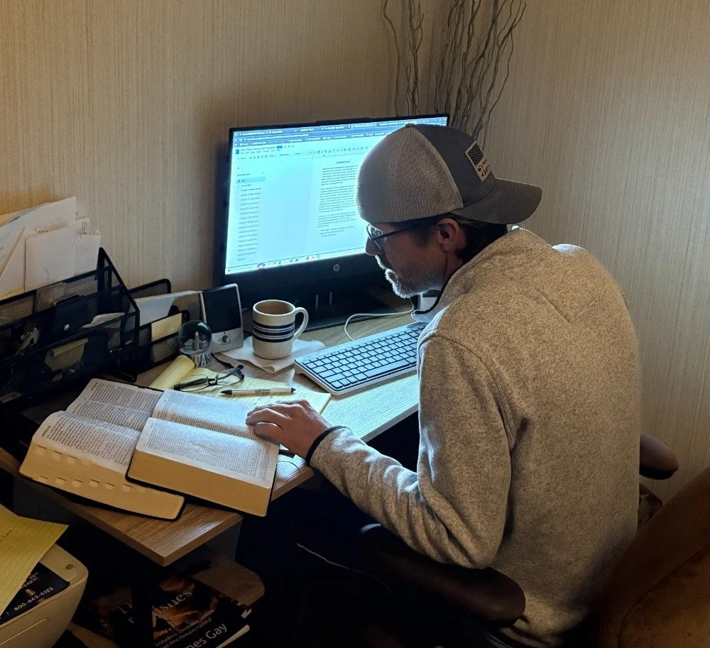 A man sitting at a cluttered desk, working on a computer, surrounded by books, a coffee mug, and office supplies.