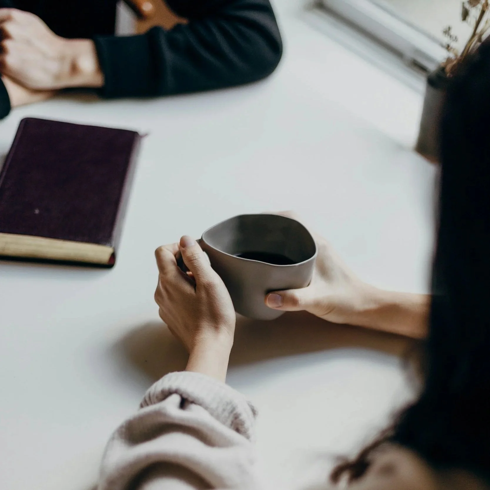 Person holding a gray coffee mug over a white table, with a closed book nearby, and another person sitting at the table.