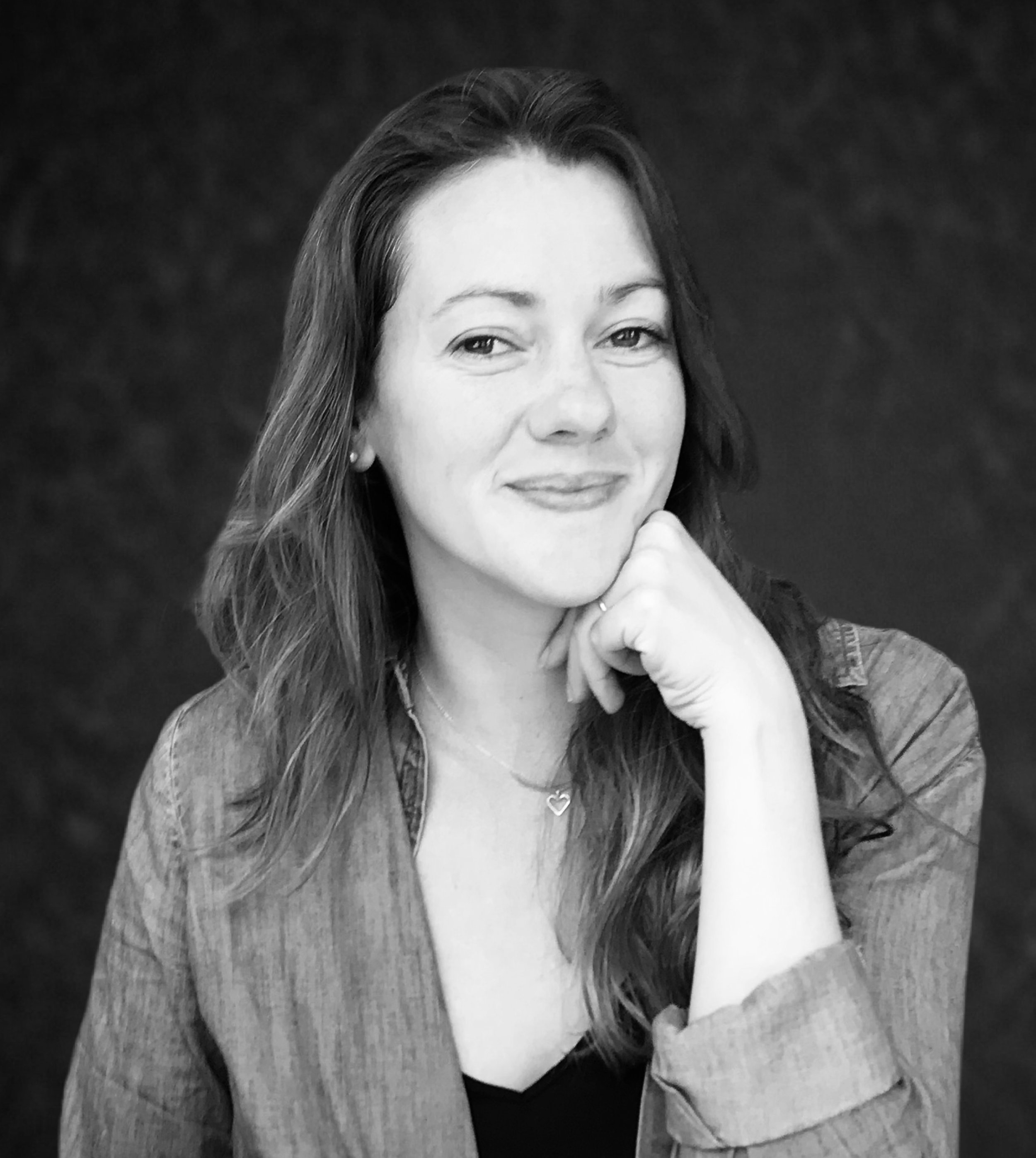 Black and white portrait of a woman with long hair, smiling, resting her chin on her left hand, wearing a jacket and a necklace with a heart pendant.