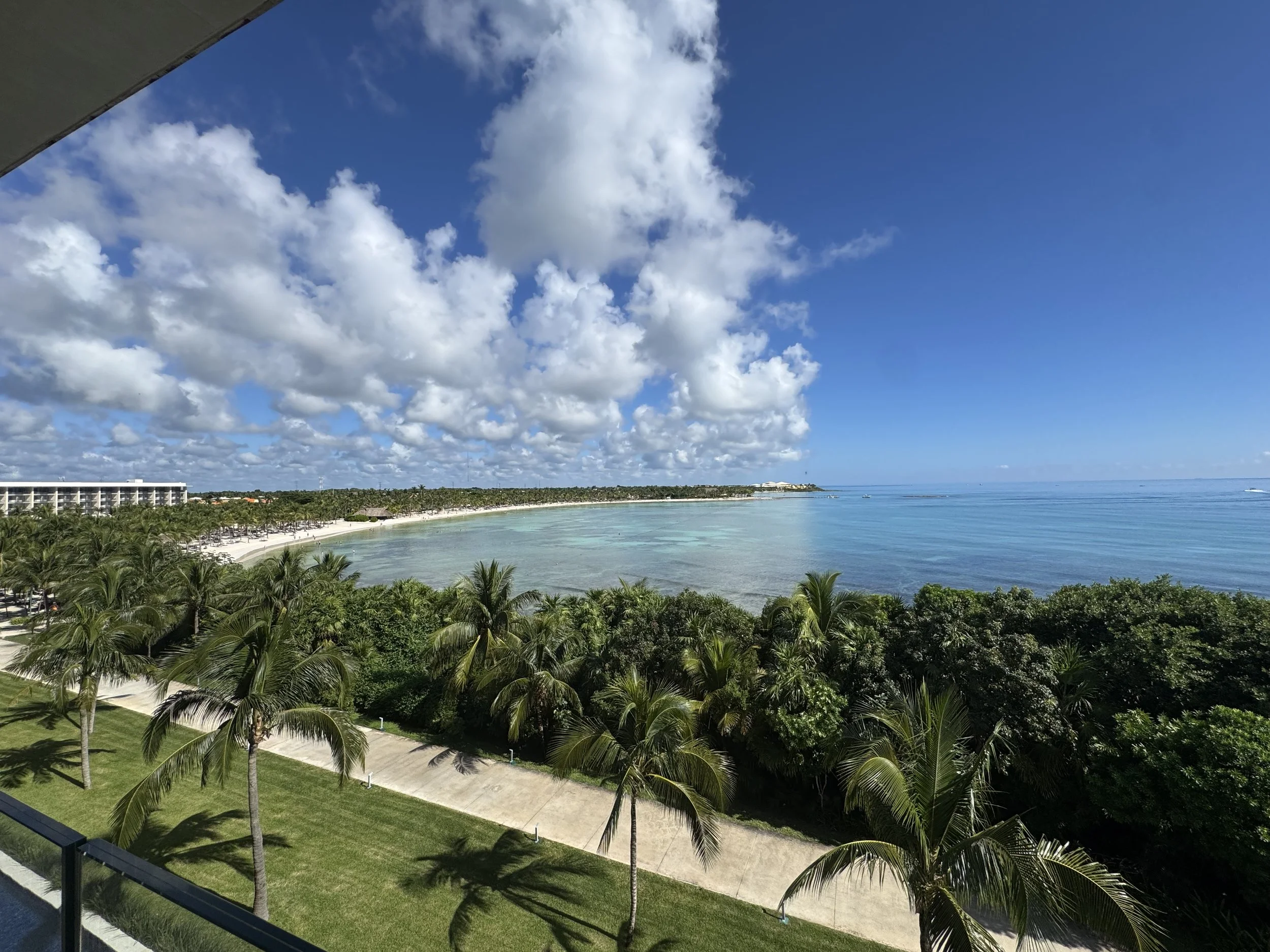 View of a beach with clear blue water, white sand, and palm trees, taken from a balcony on a sunny day with scattered clouds in the sky.