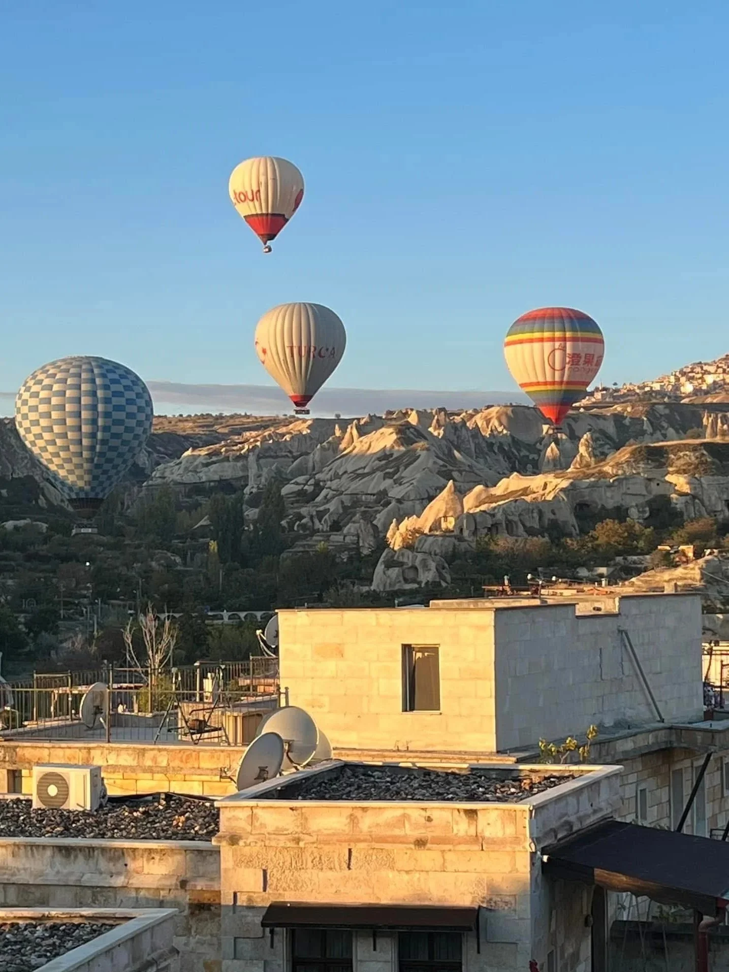 Five hot air balloons floating above rocky hills during sunrise over city buildings.