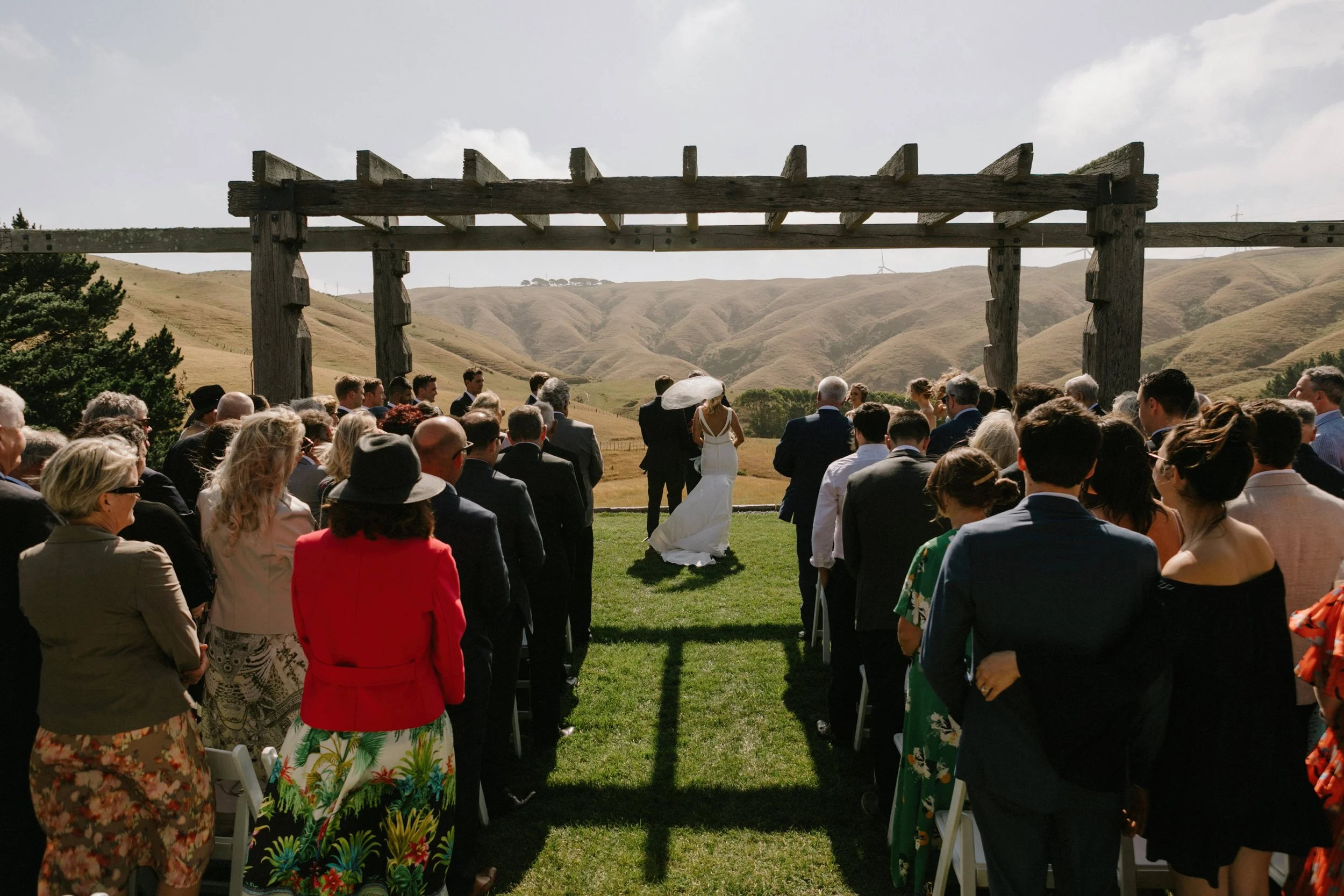 Outdoor wedding ceremony with guests seated on grass facing a bride and groom under a large wooden arch in a scenic valley surrounded by rolling hills.